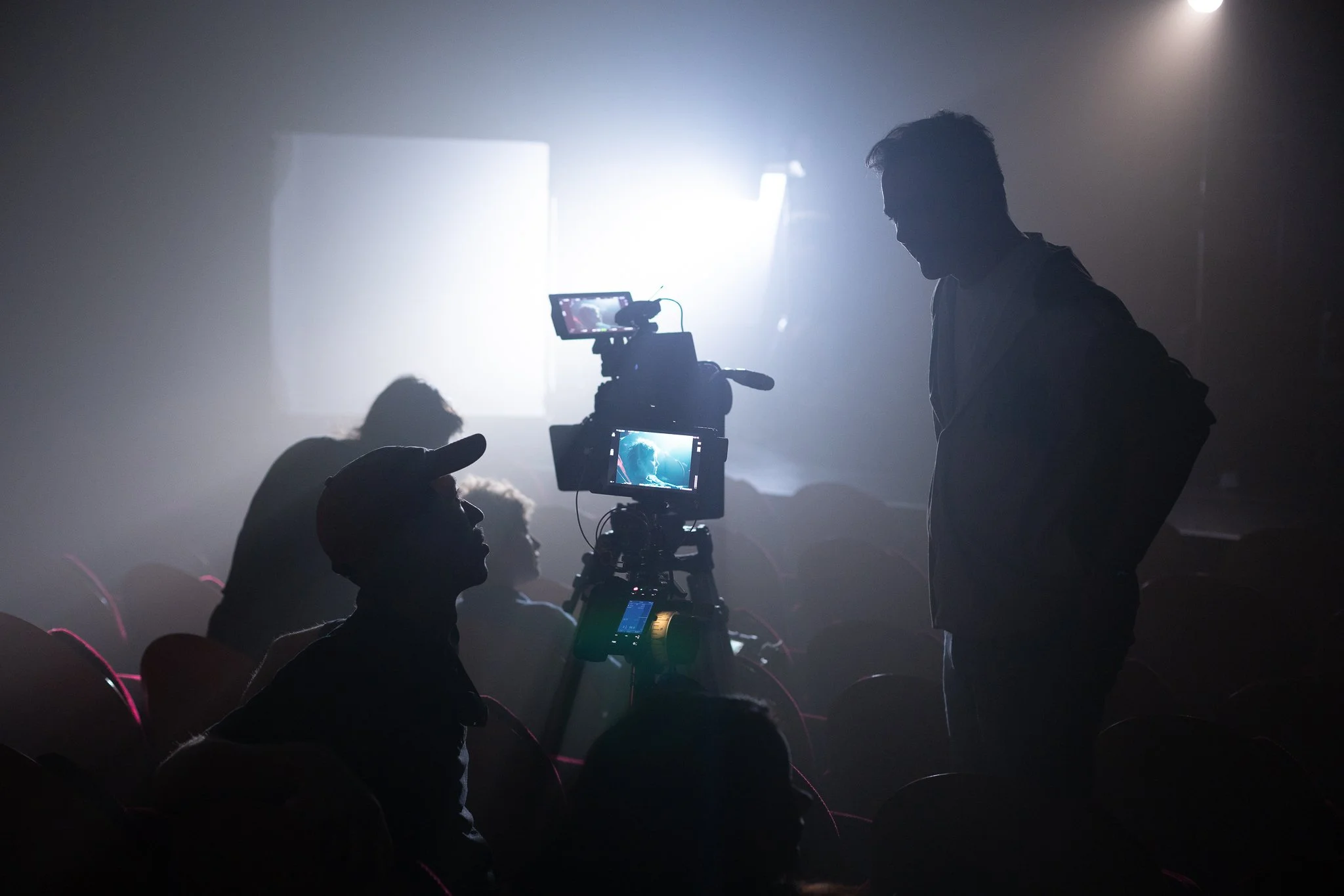 Une scène en coulisses d'une production vidéo, avec une caméra filmant un acteur ou une actrice sur scène, entourée de membres de l'équipe technique dans une ambiance sombre et lumineuse