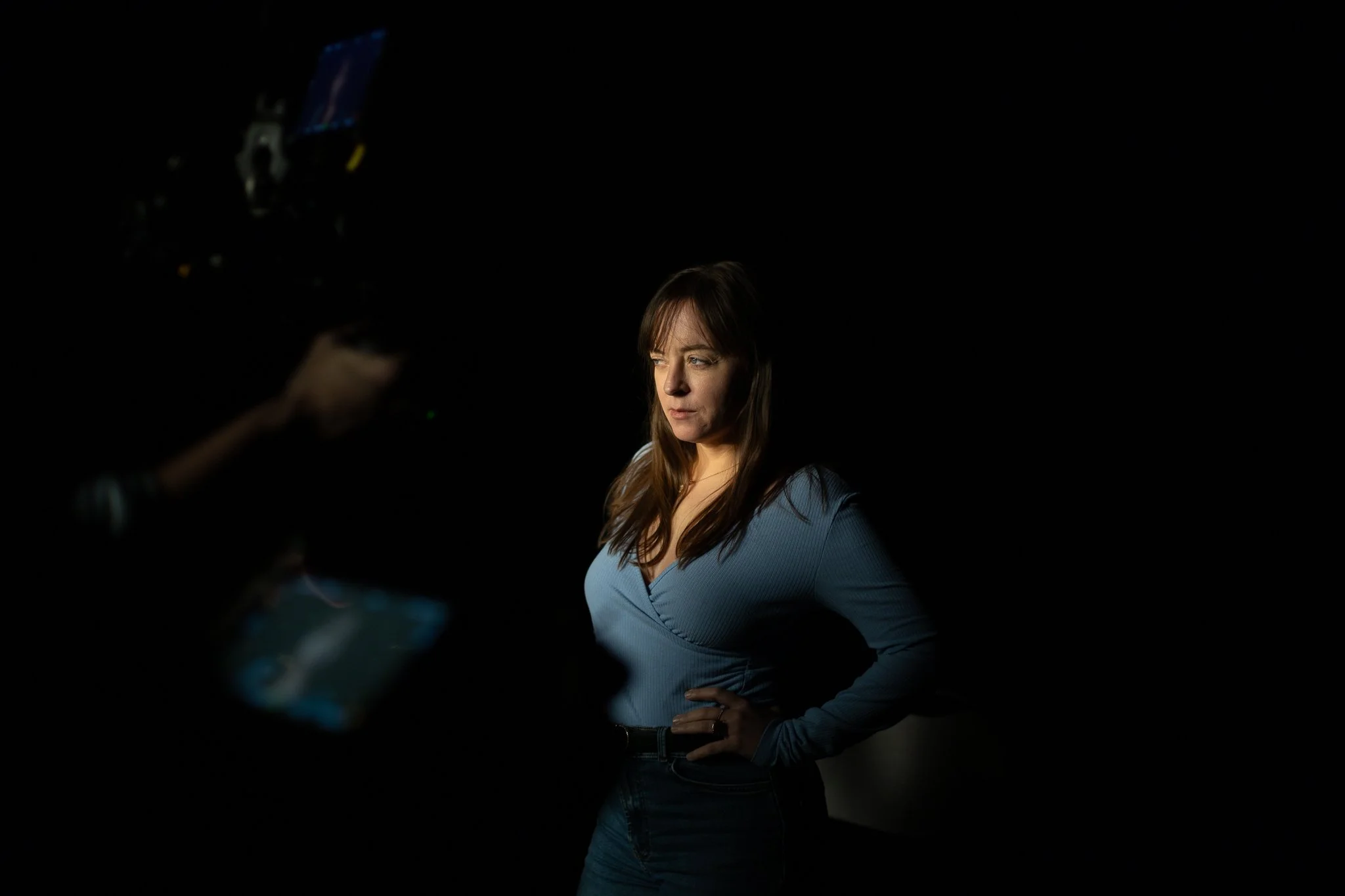 Une femme avec des cheveux bruns, portant un haut bleu, pose pour un shooting photo dans un environnement sombre avec un éclairage ciblé.