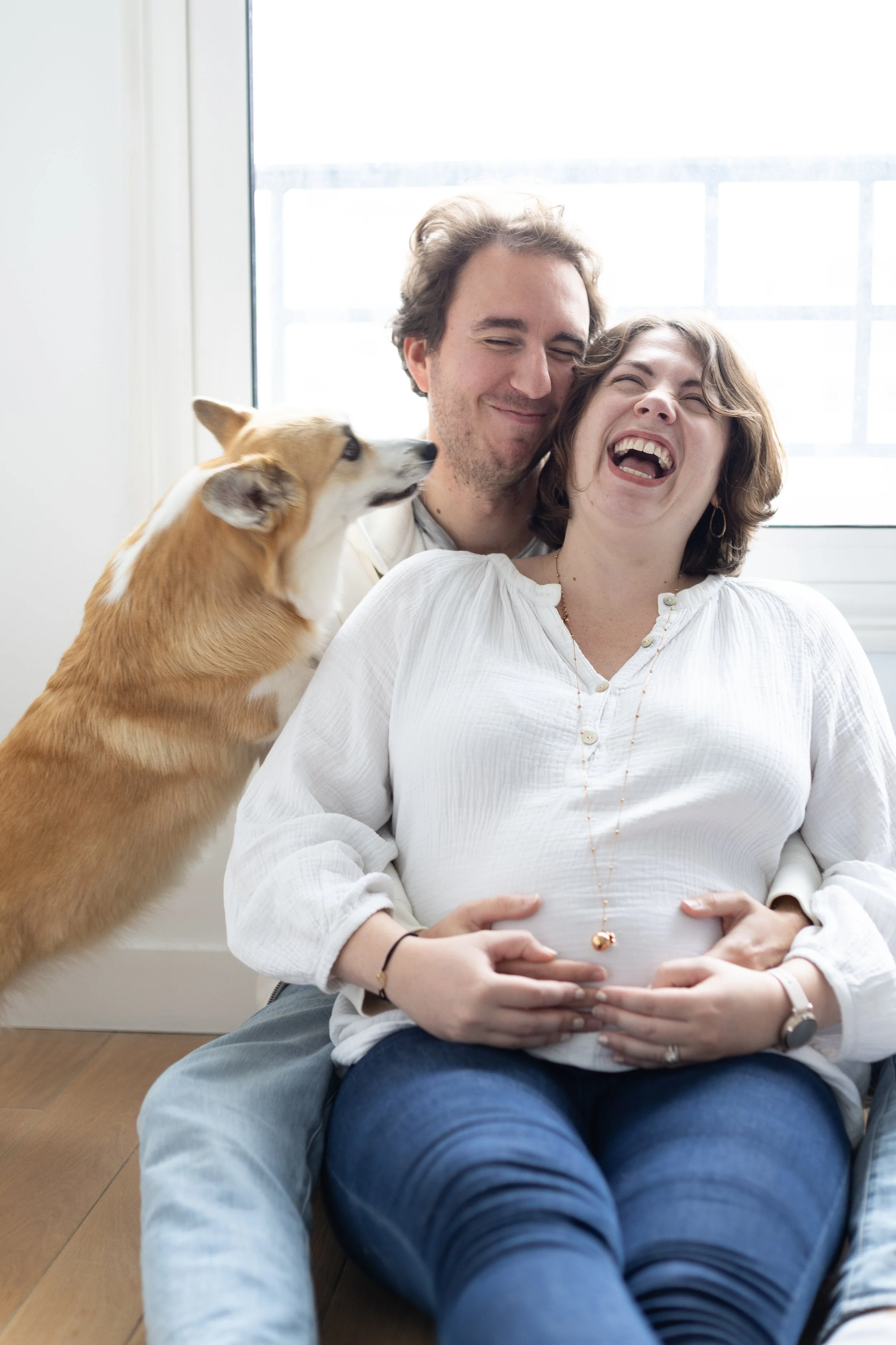 Un couple souriant avec un chien, la femme enceinte, près d'une grande fenêtre lumineuse.