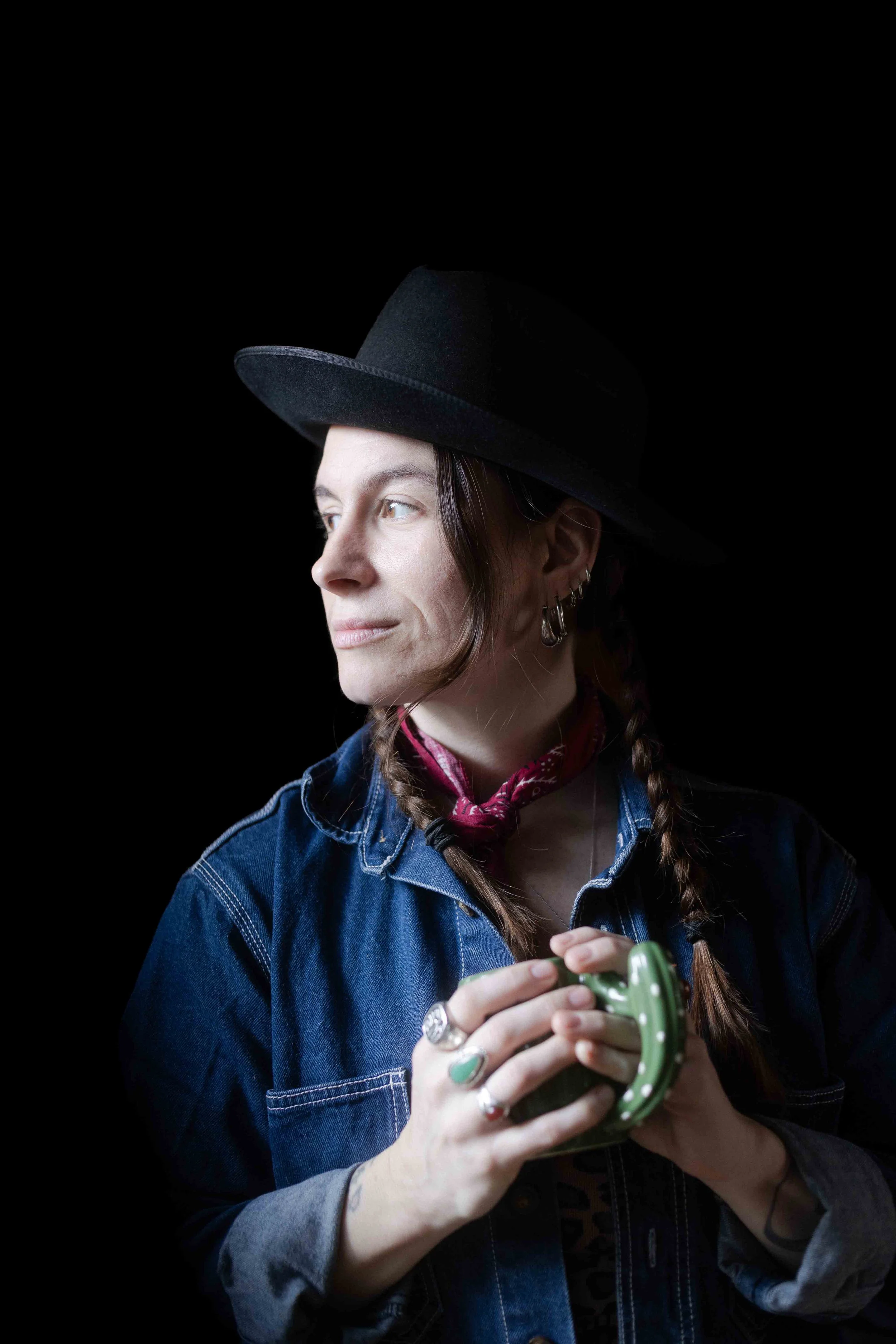 Une jeune femme portant un chapeau noir, une veste en jean et une bandana rouge, tenant une cuillère ou un ustensile en forme de fer à cheval vert avec des pois blancs, contre un fond noir.