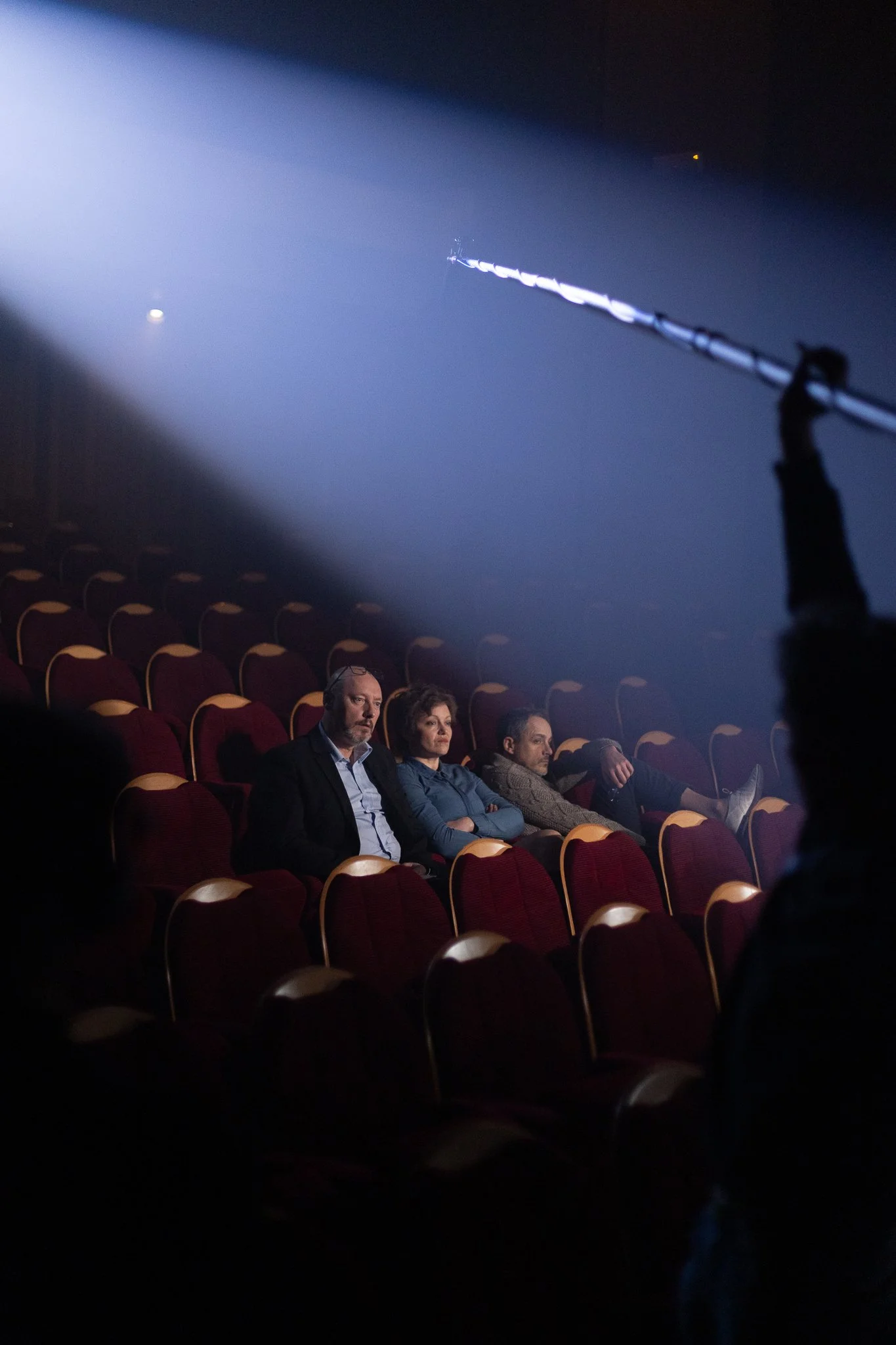 Une salle de cinéma avec des spectateurs assis, regardant un écran, éclairée par la lumière d'un projecteur ou d'une scène. Présence de quatre personnes à l'avant, concentrées sur l'écran.