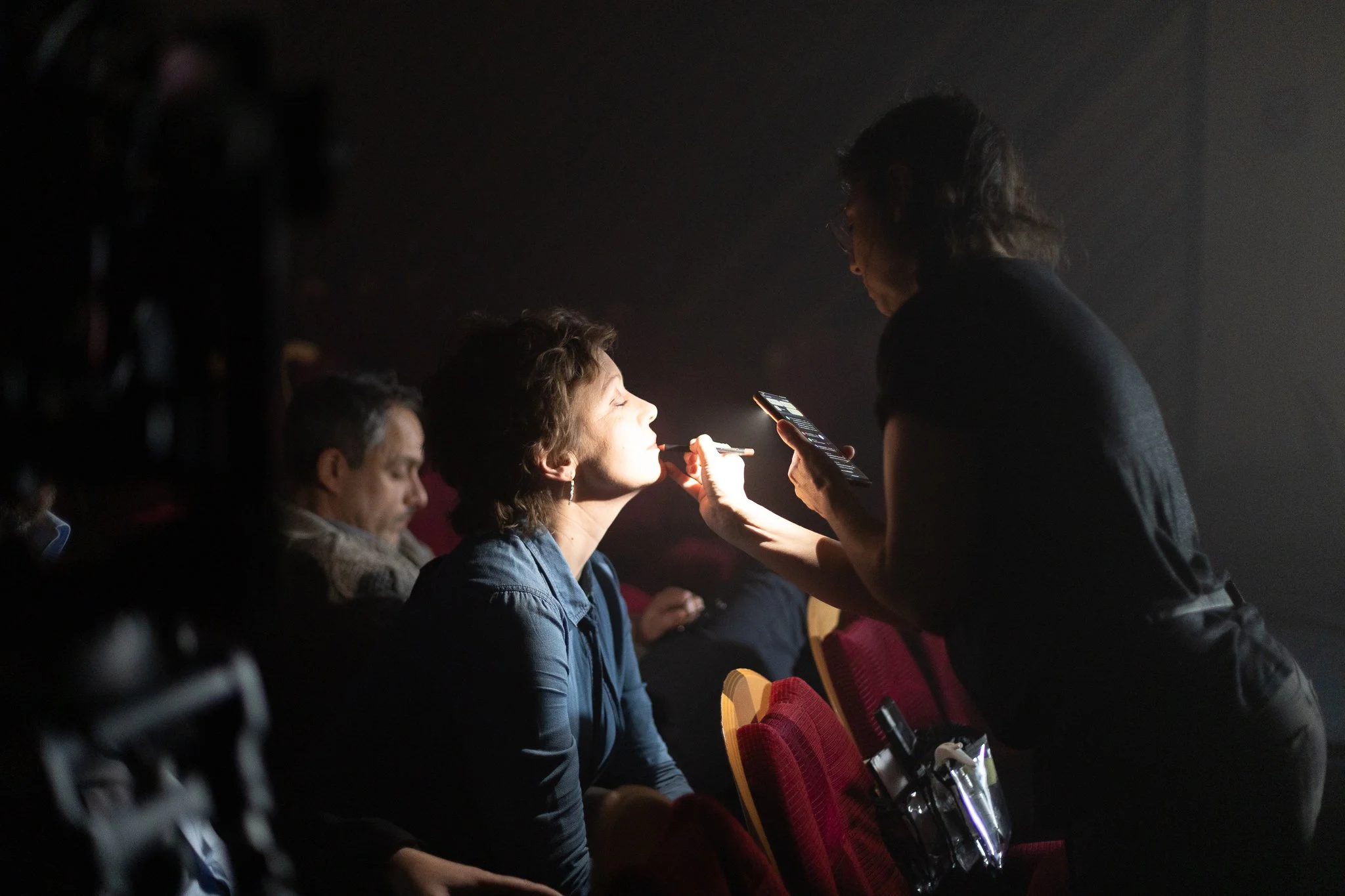 Une femme maquille une autre femme dans une salle sombre, probablement lors d'une séance de maquillage ou d'un atelier.