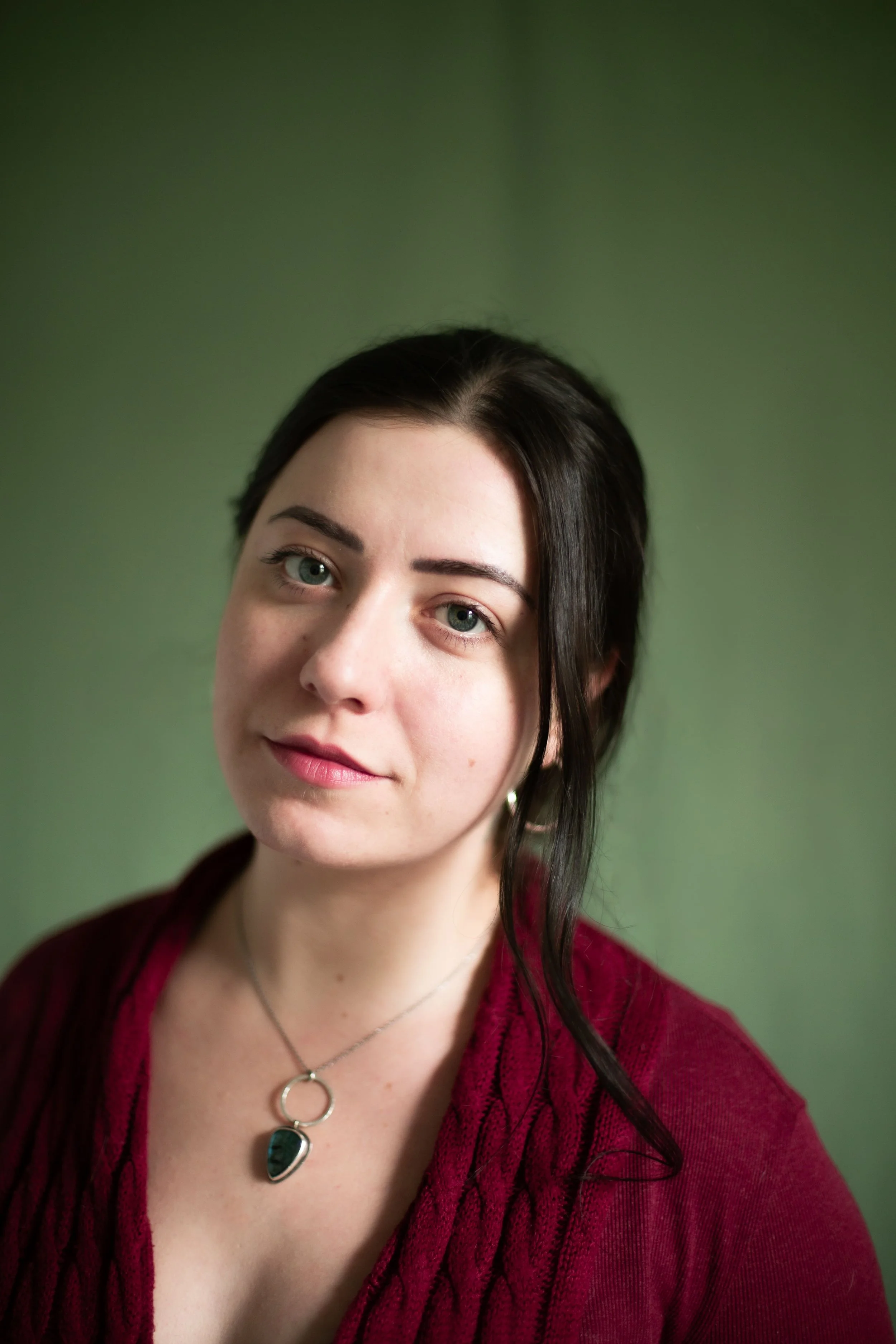Portrait d'une femme aux cheveux noirs, yeux verts, portant un chemisier rouge et un collier avec un pendentif en forme de goutte, contre un fond vert