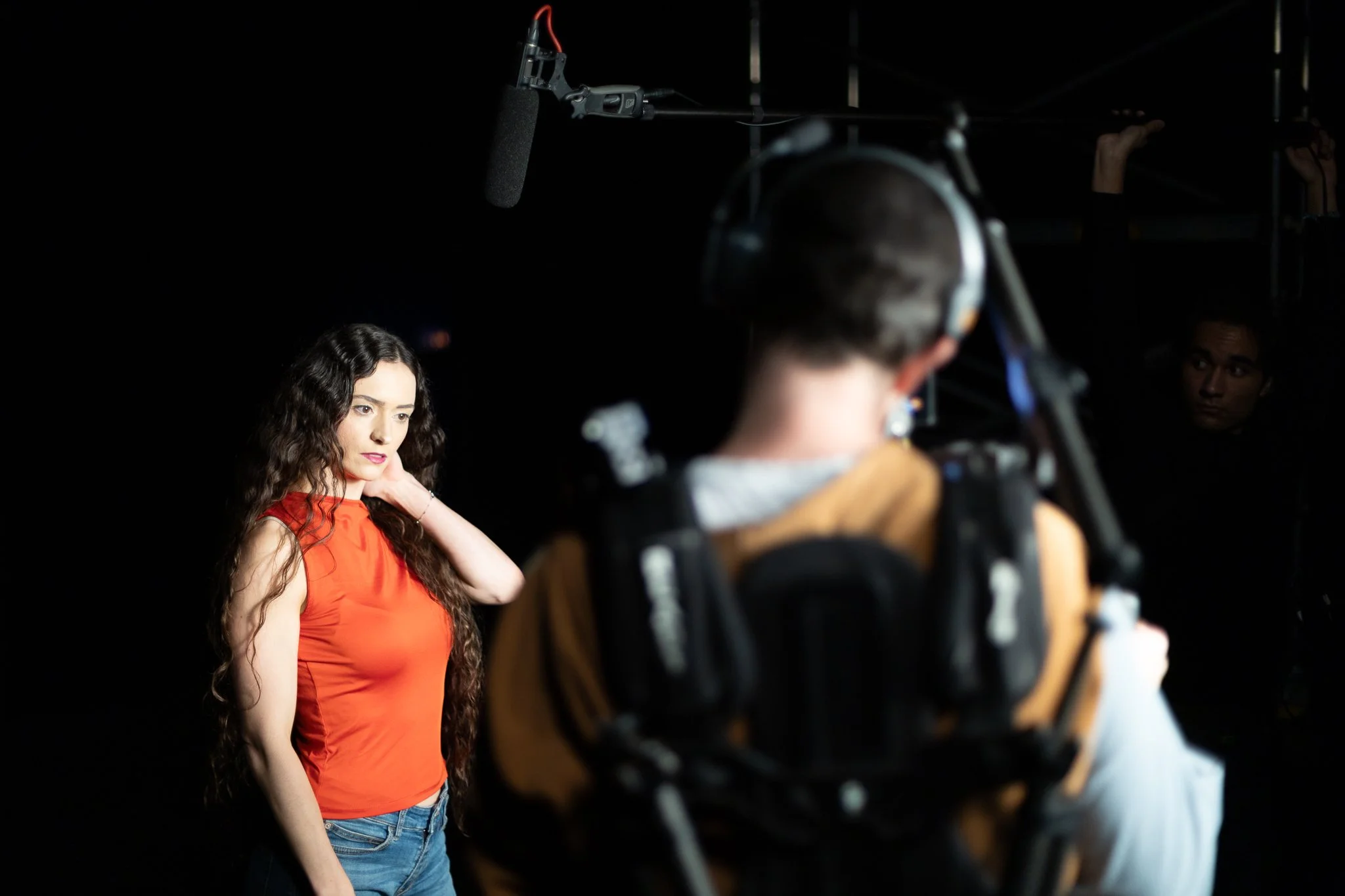 Une femme avec des cheveux ondulés, portant un débardeur rouge, se prépare pour une séance de tournage avec un microphone et un caméraman