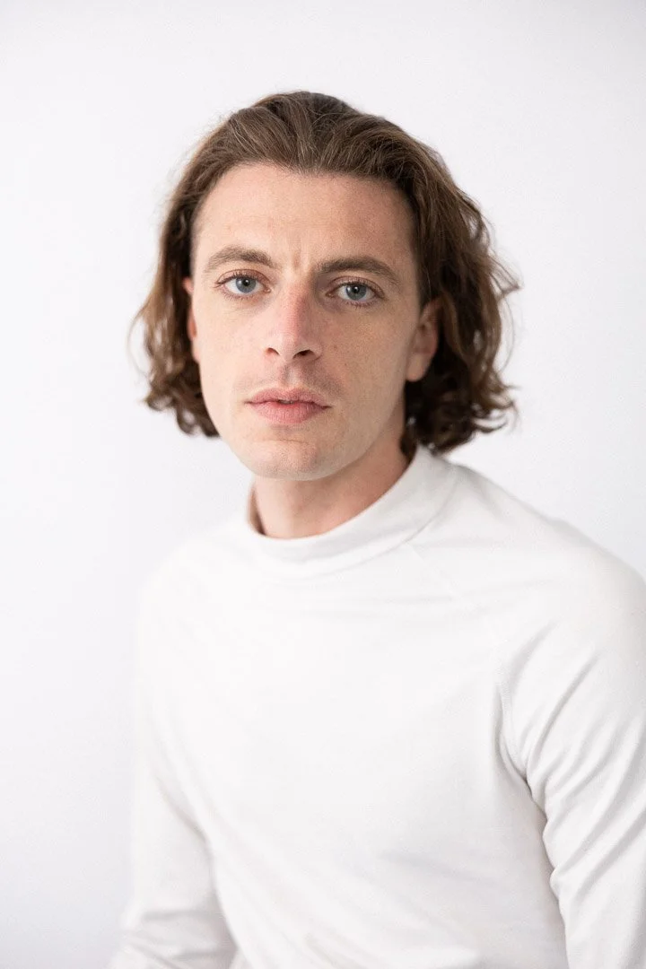 Un jeune homme avec des cheveux bouclés et blonds, portant un t-shirt blanc, regarde l'objectif avec une expression neutre, contre un fond blanc.