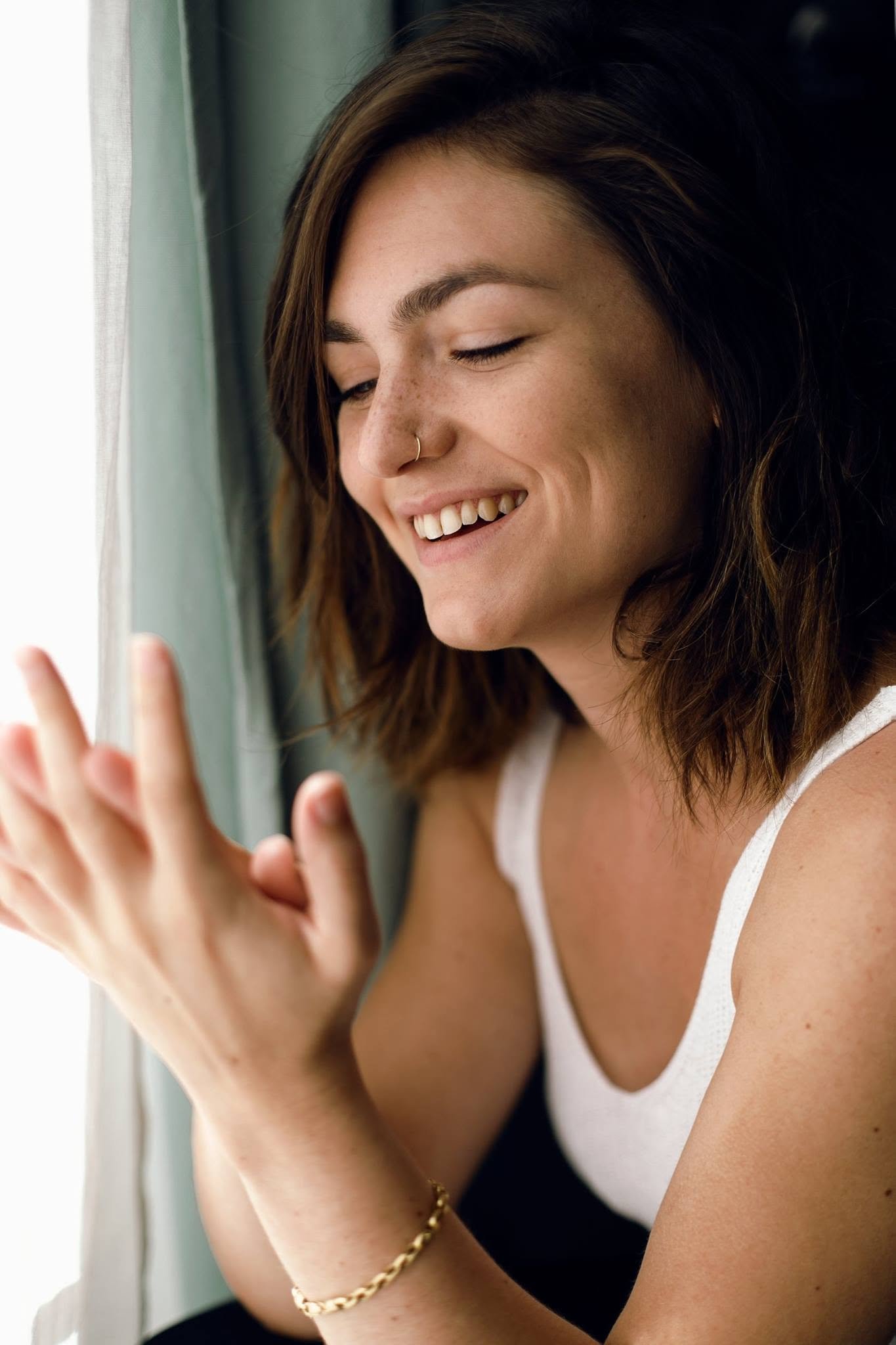 Une femme avec des cheveux bruns, souriant, regarde par une fenêtre et tient sa main levée devant la vitre, portant un bracelet en or.
