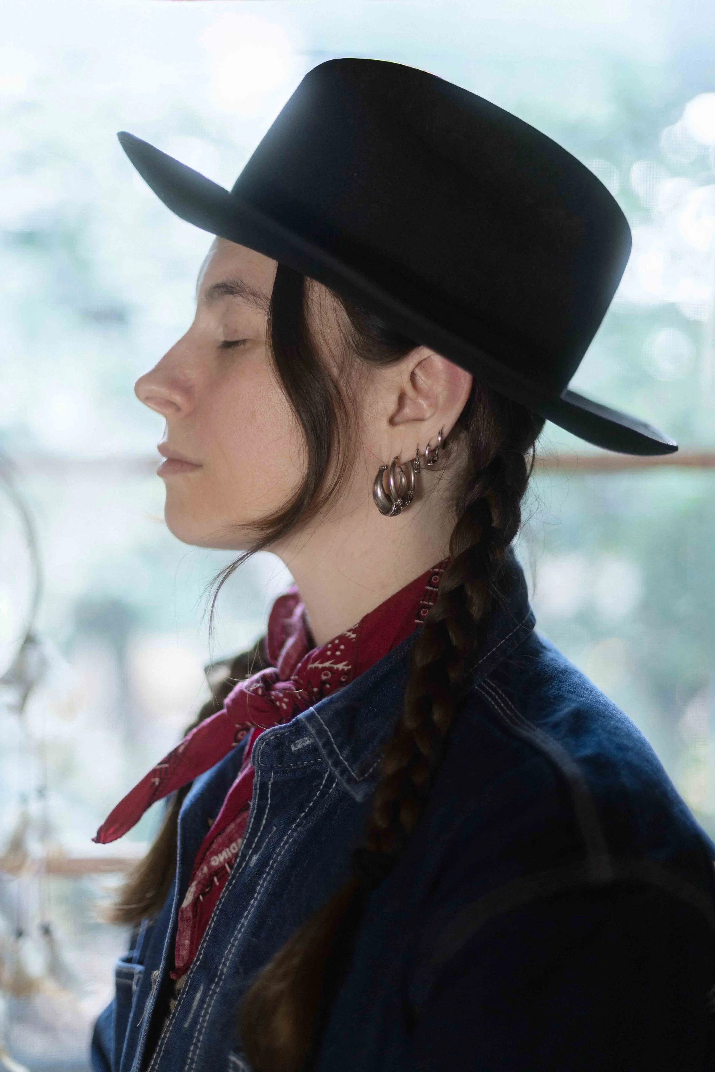 Jeune femme avec une tresse, portant une veste en jean, une chemise rouge et un bandana rouge, portant plusieurs piercings à l'oreille, portant un grand chapeau noir, pause en intérieur.