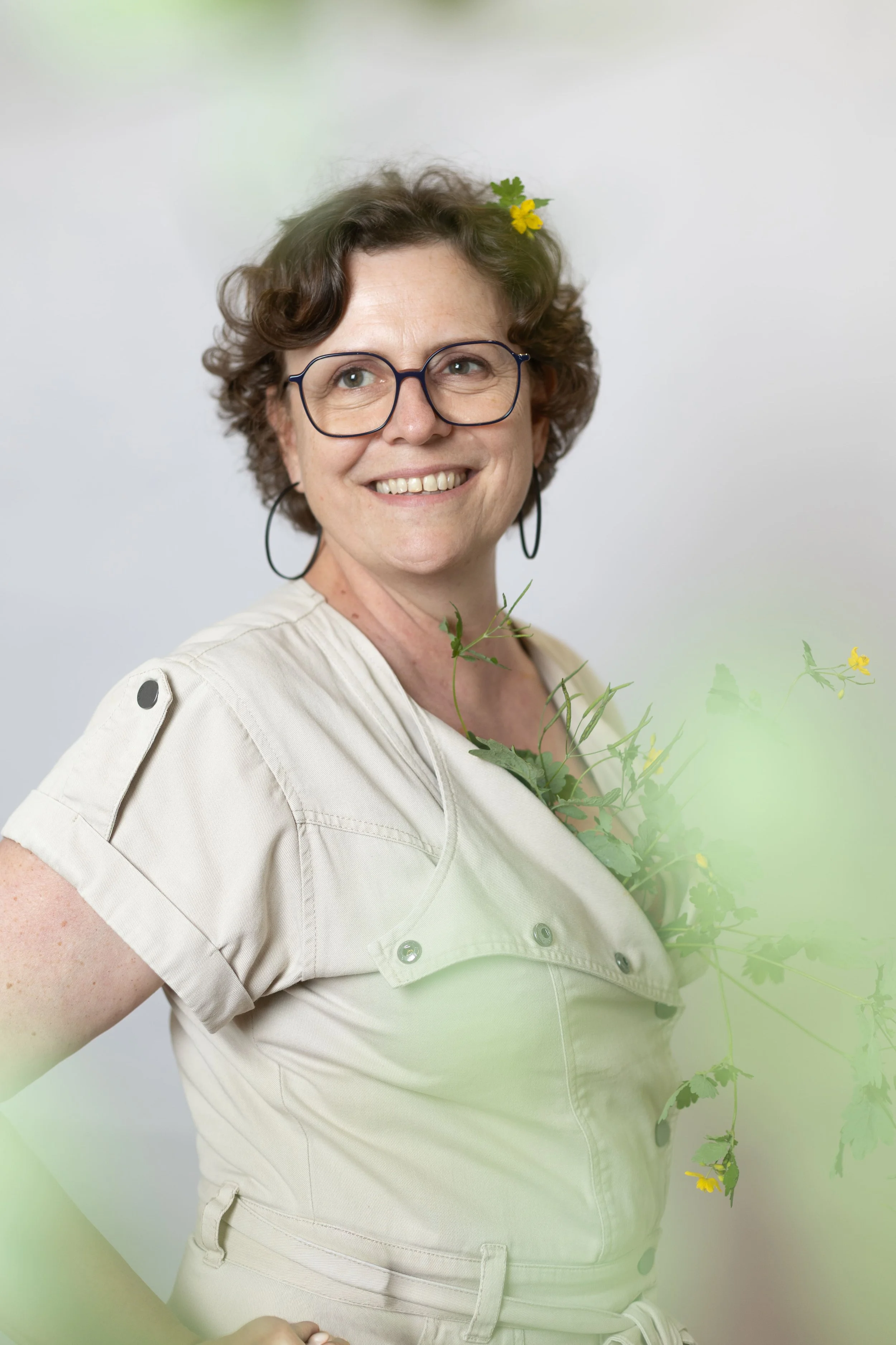 Une femme souriante portant des lunettes, avec des fleurs jaunes dans ses cheveux et des feuilles vertes, portant une tenue beige, en arrière-plan blanc.