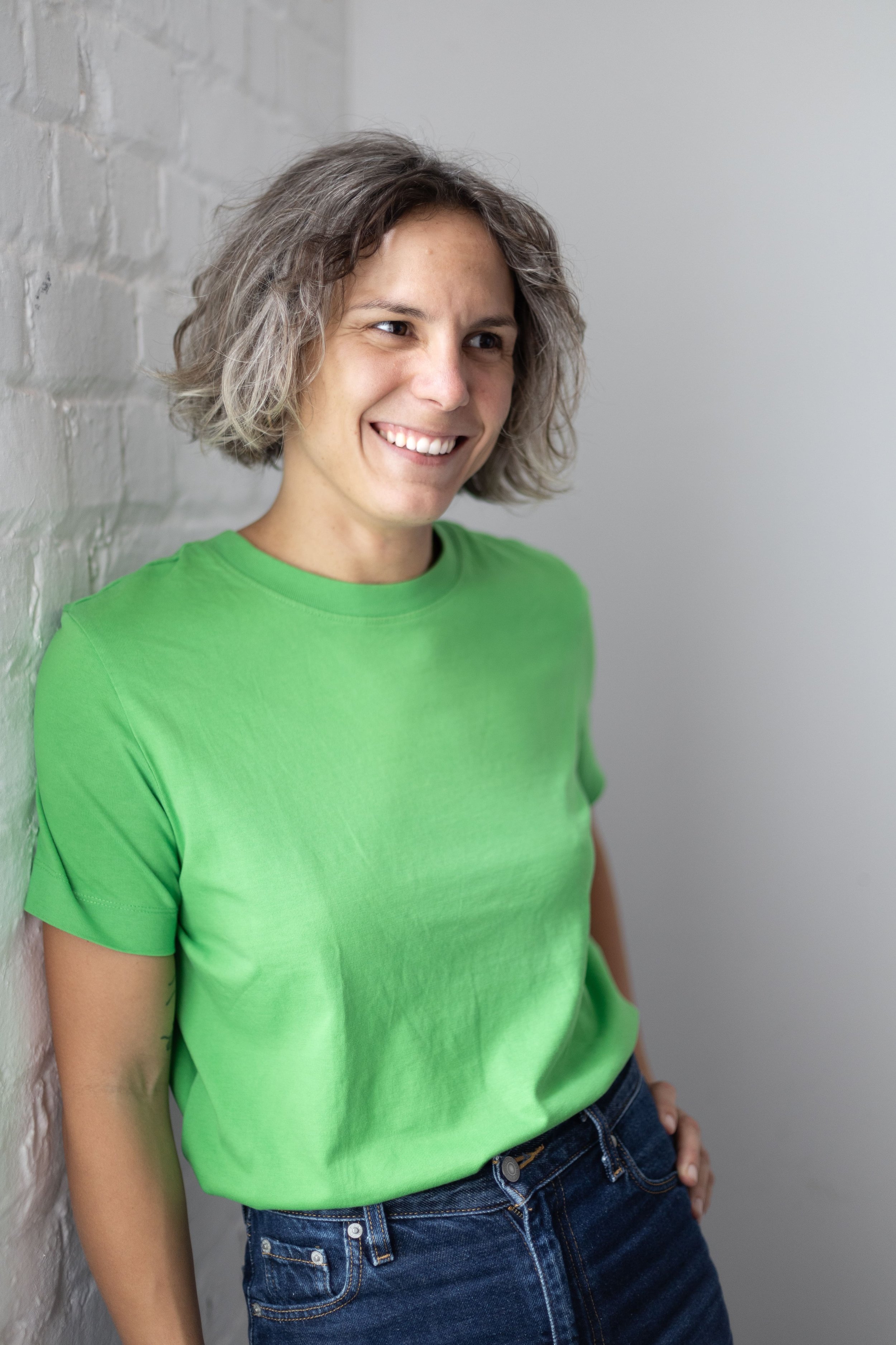 Une femme souriante, portant un t-shirt vert vif et un jean, debout contre un mur blanc. Elle a les cheveux courts, légèrement bouclés, et regarde vers la gauche.