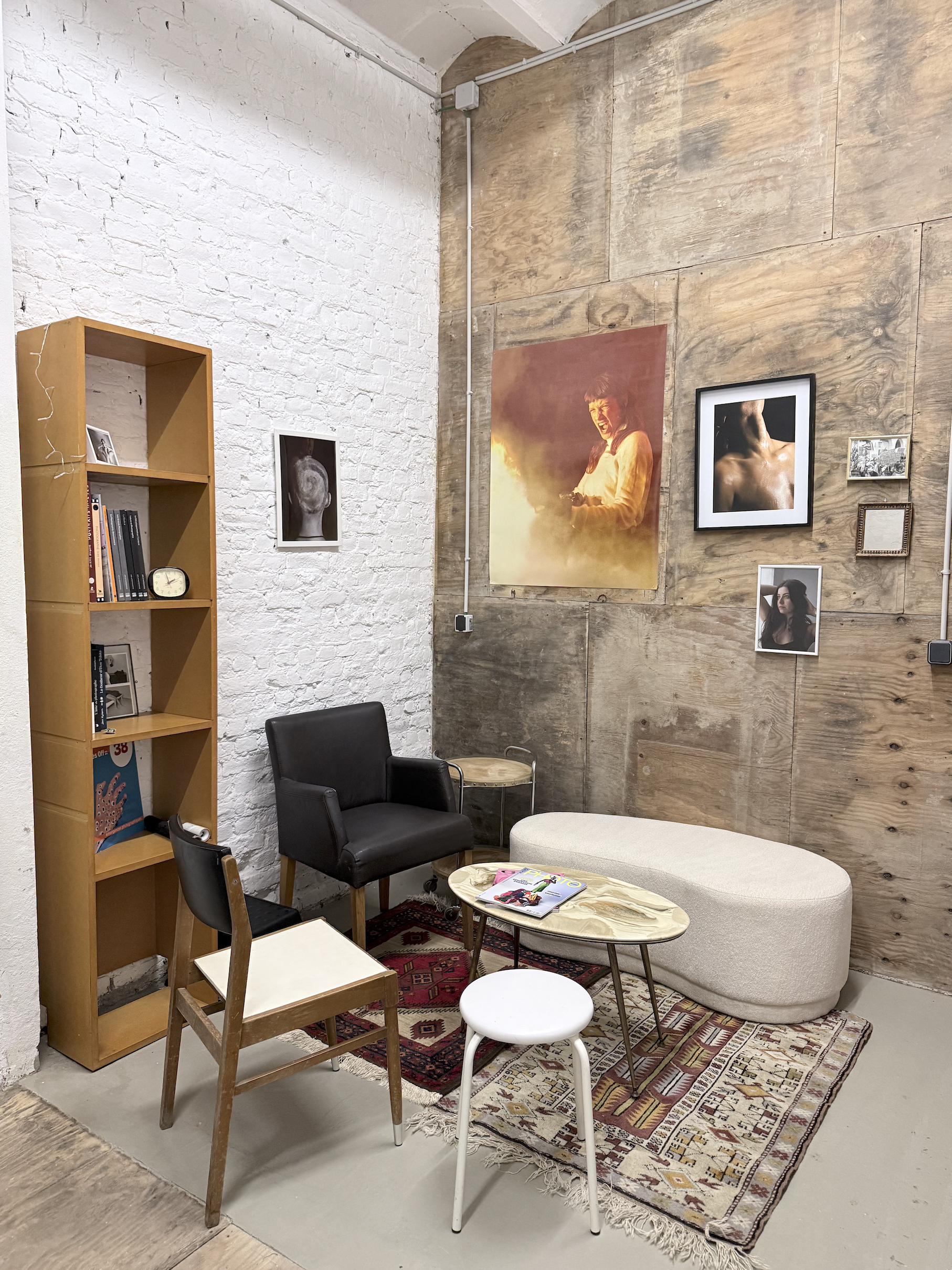Une salle de séjour avec un mur de briques blanches et un mur en bois avec des cadres photo. Il y a un canapé beige, deux chaises noires, un petit tapis coloré, une table ronde avec des magazines, un tabouret blanc, une étagère en bois avec des livres et des cadres. Des œuvres d'art sont accrochées au mur.
