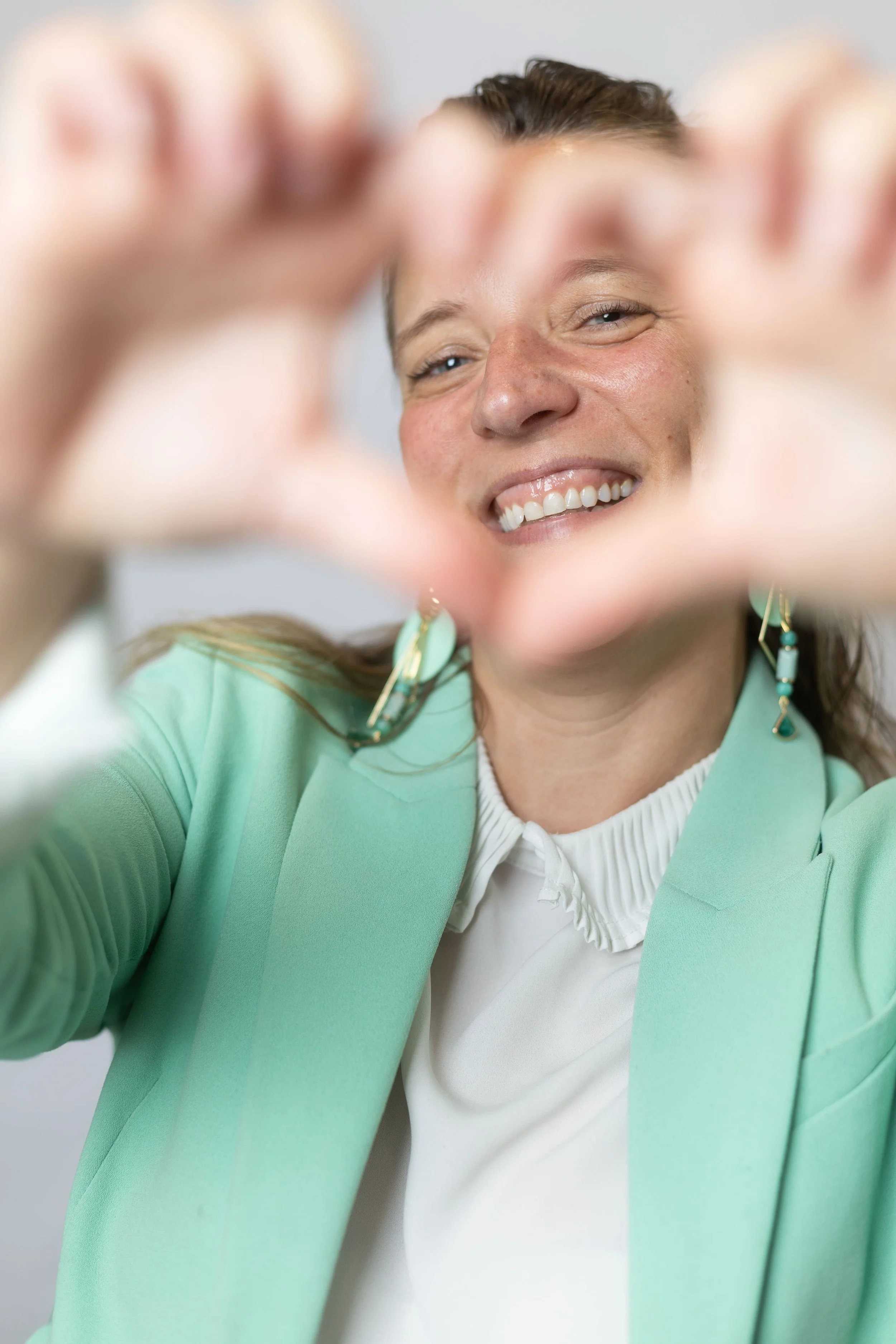 Une femme souriante qui prend un selfie, portant une veste verte et une chemise blanche avec des boucles d'oreilles turquoise.