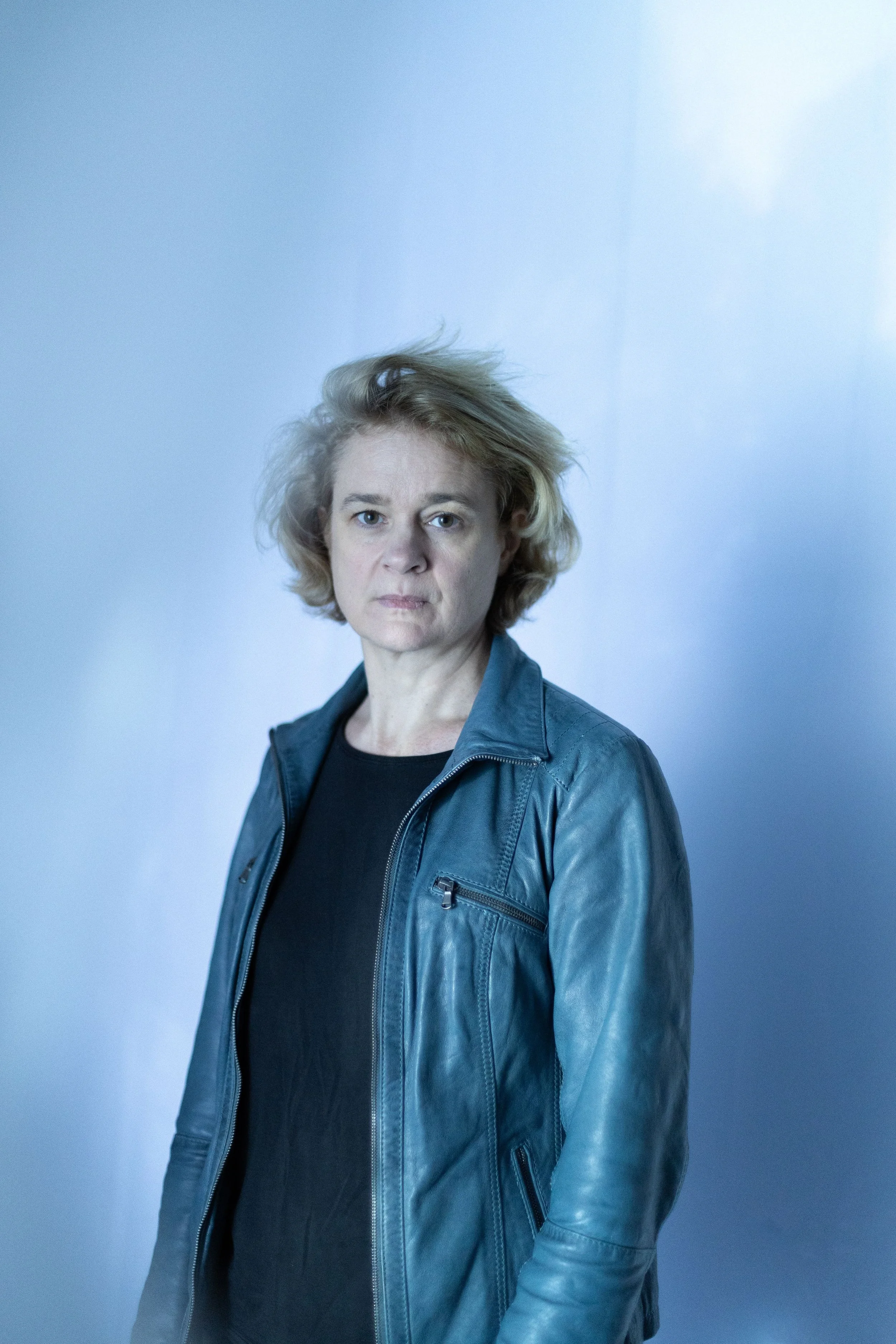 Portrait d'une femme avec des cheveux blonds, portant une veste en cuir gris et un t-shirt noir, se tenant devant un mur blanc avec un éclairage doux.