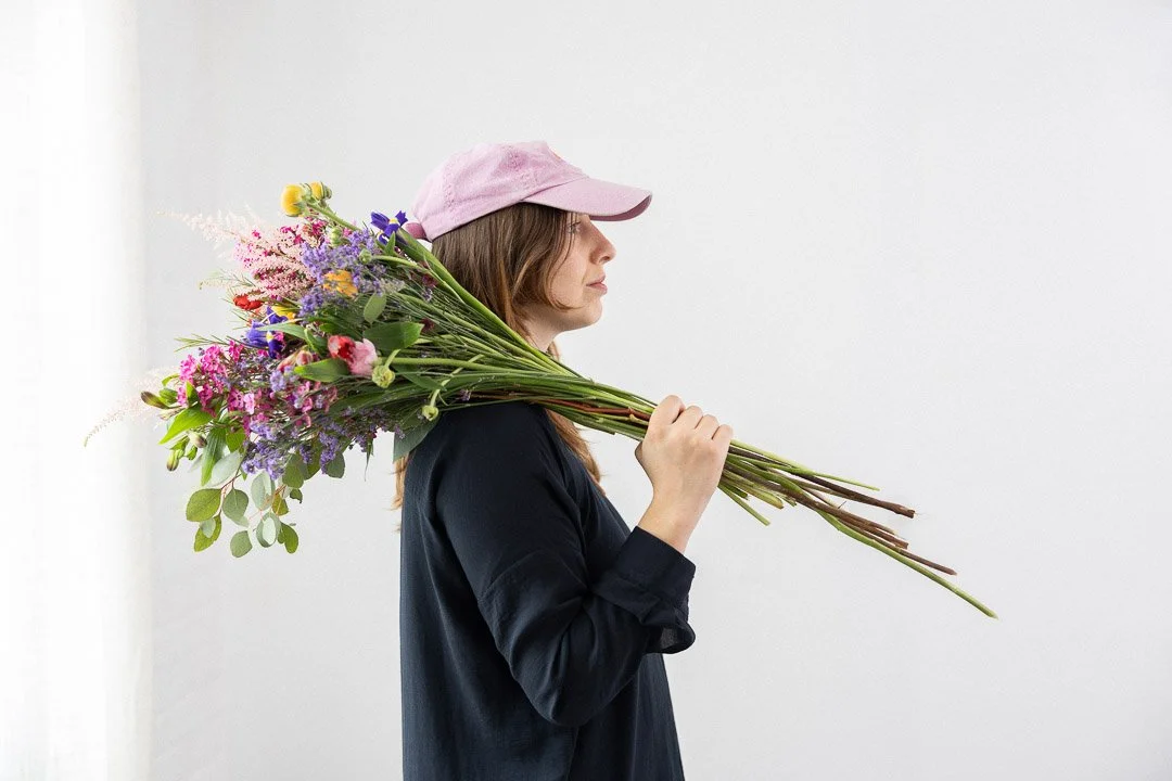 Femme portant une casquette rose et une chemise sombre, tenant un bouquet de fleurs colorées sur l'épaule.