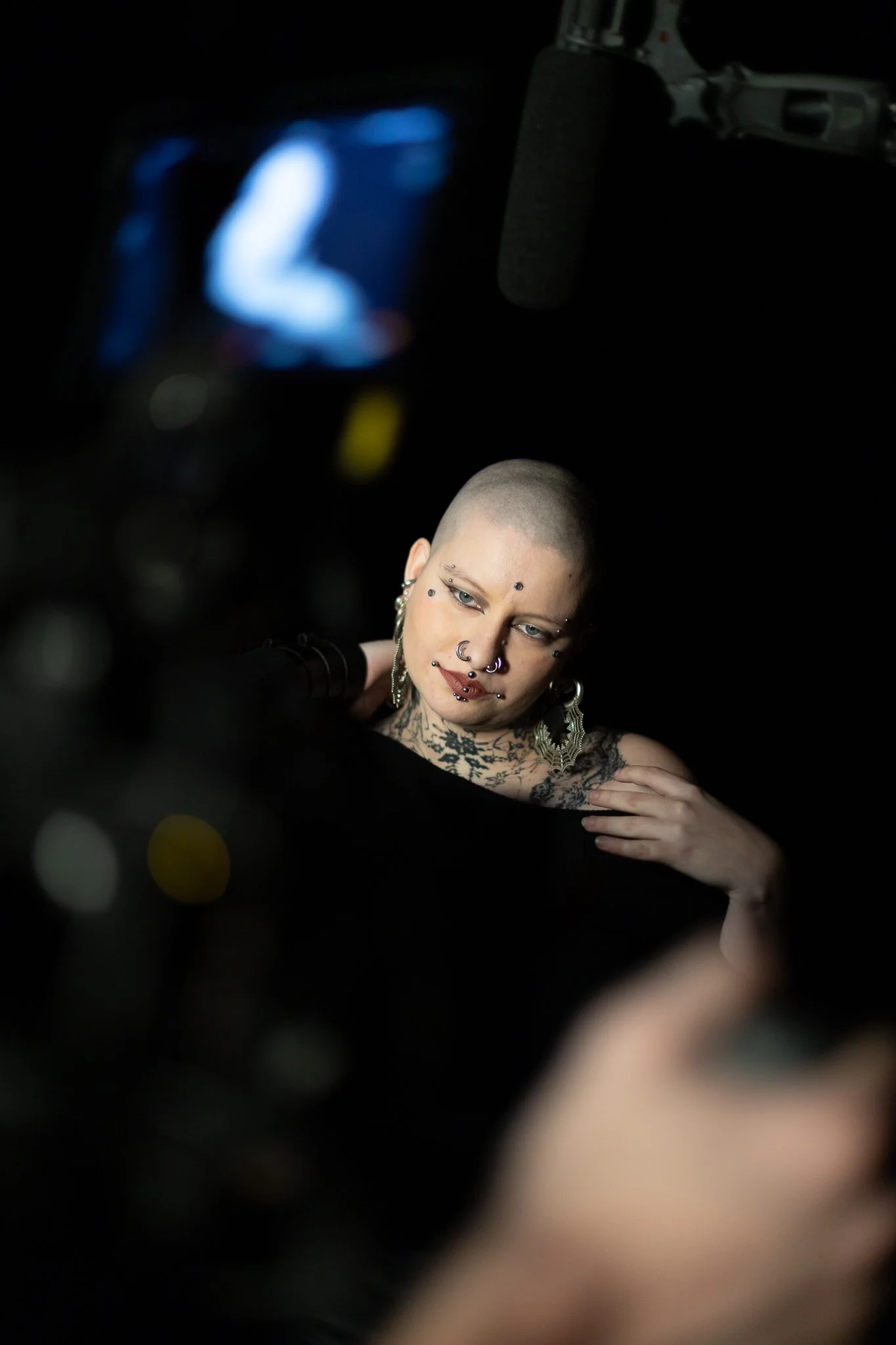 Un portrait d'une femme avec une tête rasée, portant plusieurs piercings et des boucles d'oreilles en forme de cheveux. Elle a des tatouages visibles sur son épaule et son cou, et regarde vers le bas avec une expression sérieuse. L'arrière-plan est s