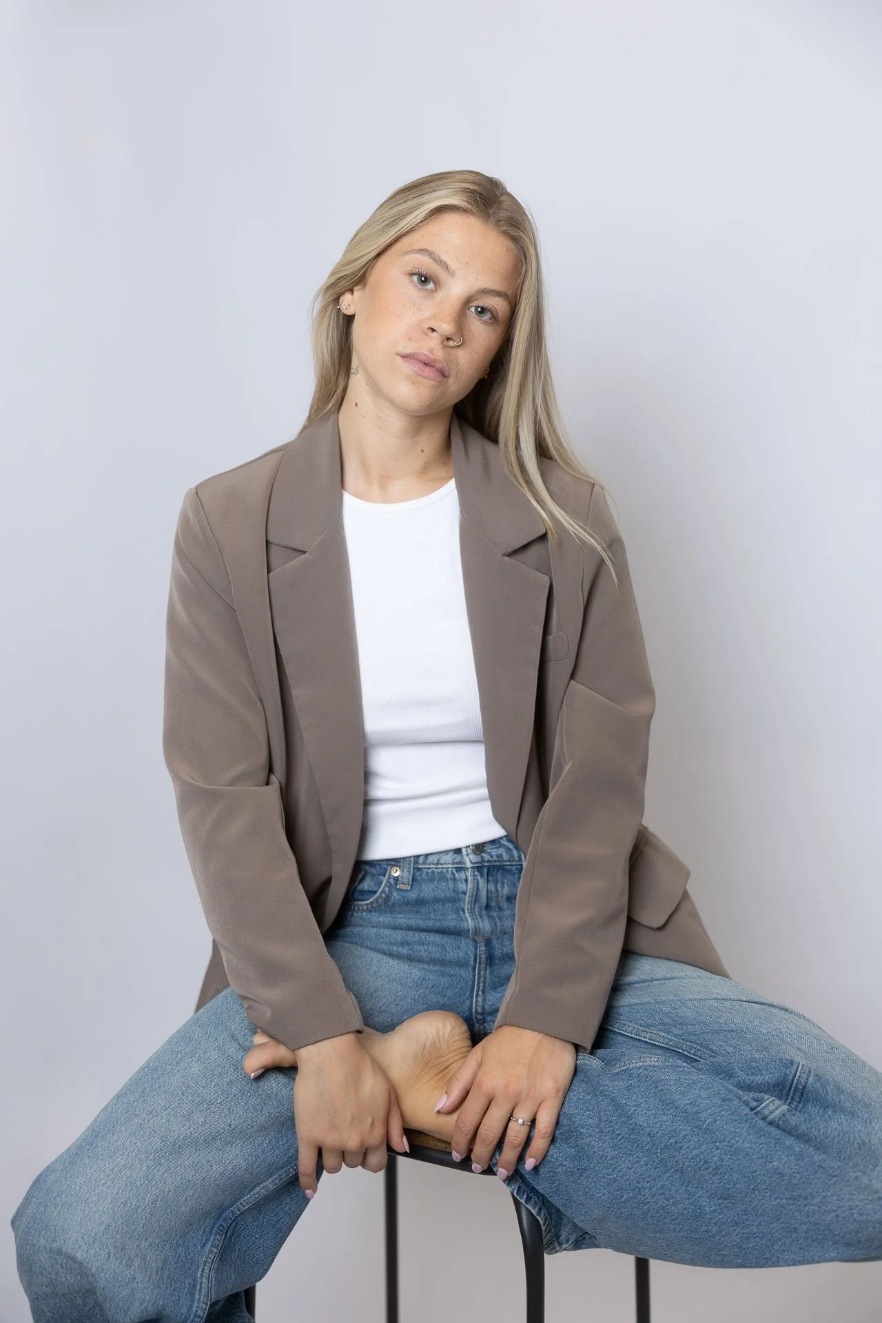 Jeune femme aux cheveux blonds, portant un blazer beige, un t-shirt blanc, et un jean bleu, assise sur une chaise avec une expression neutre, tenant son pied avec ses mains.