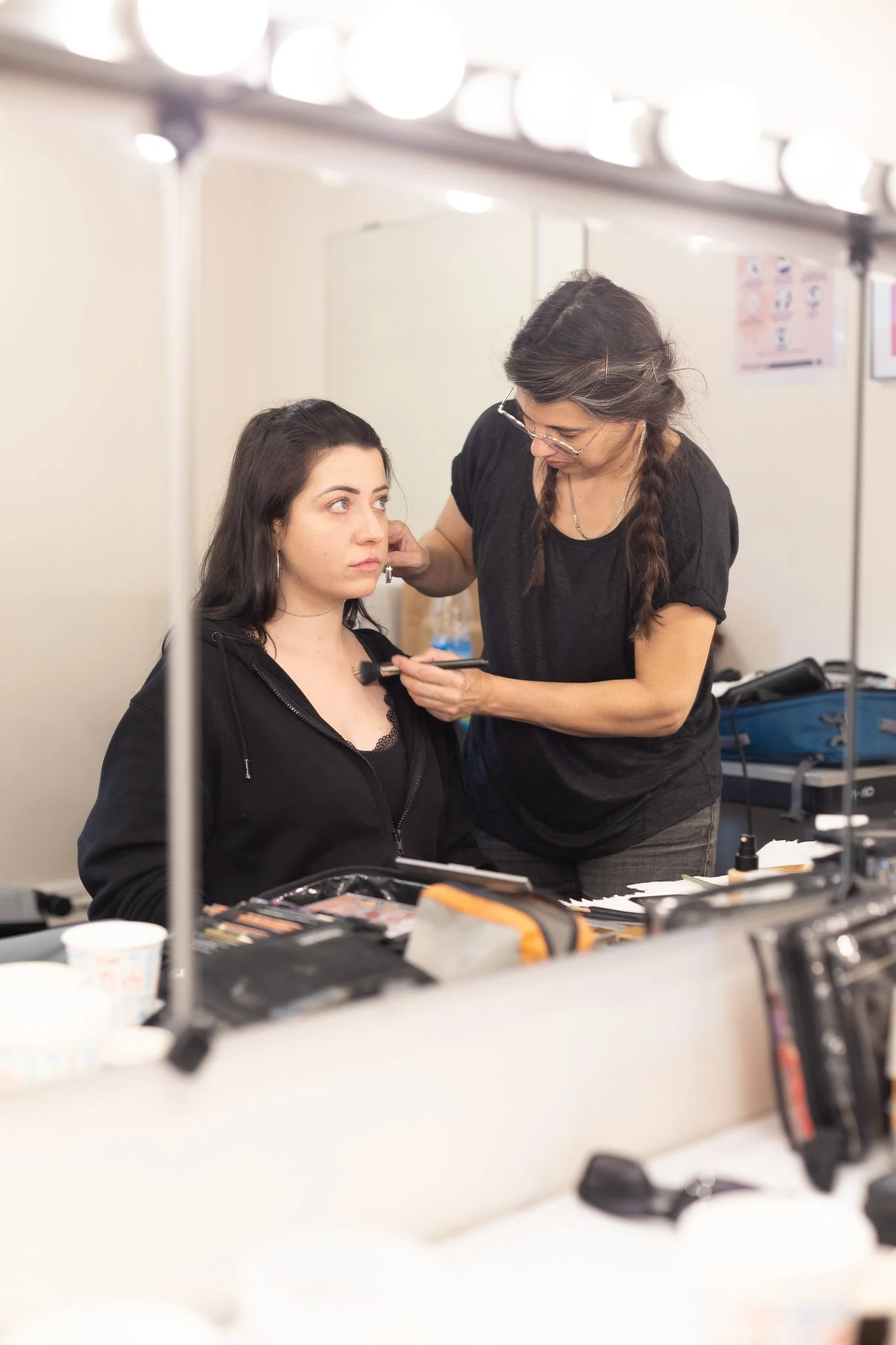 Une femme maquille une autre femme dans une cabine de maquillage, avec des lampes éclairant la scène.