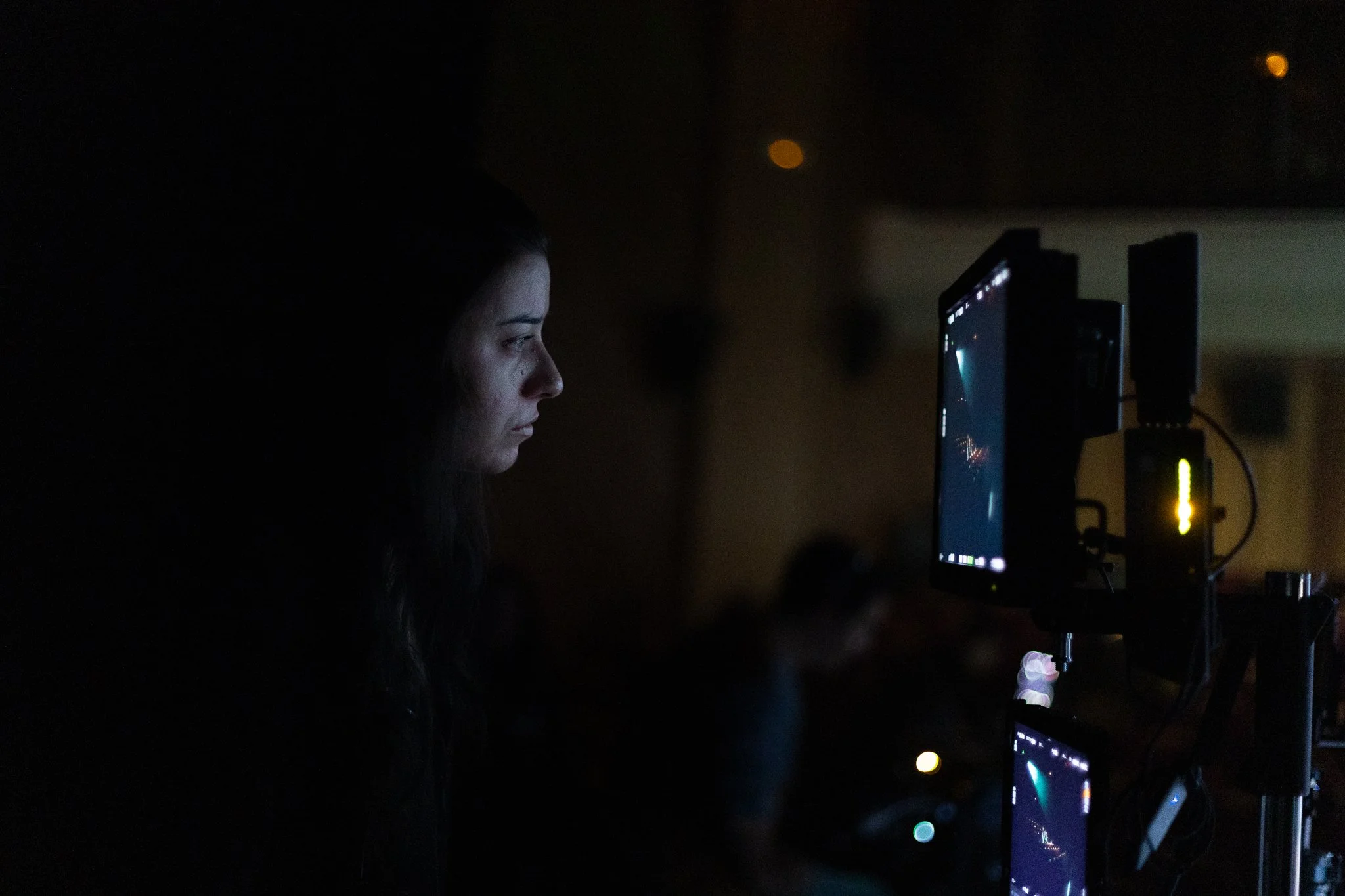 Une femme regarde un écran d'ordinateur dans une pièce sombre, avec des dispositifs électroniques allumés.