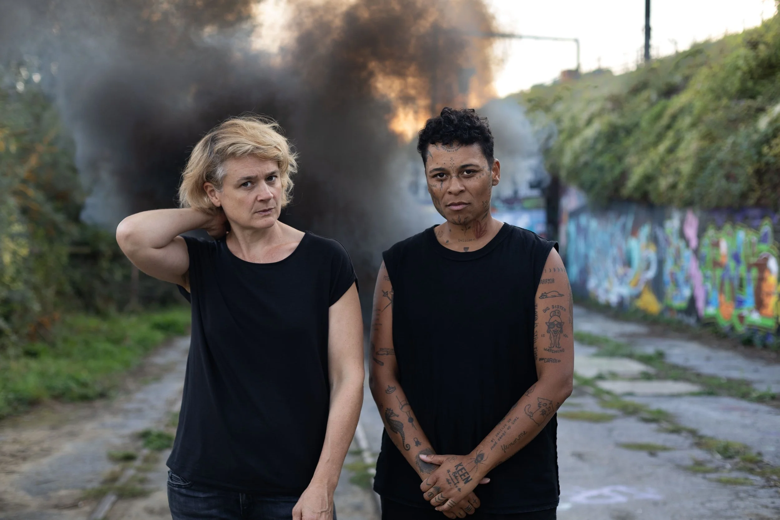Deux femmes debout sur un chemin, devant un feu et de la fumée, avec un mur graffite en arrière-plan.