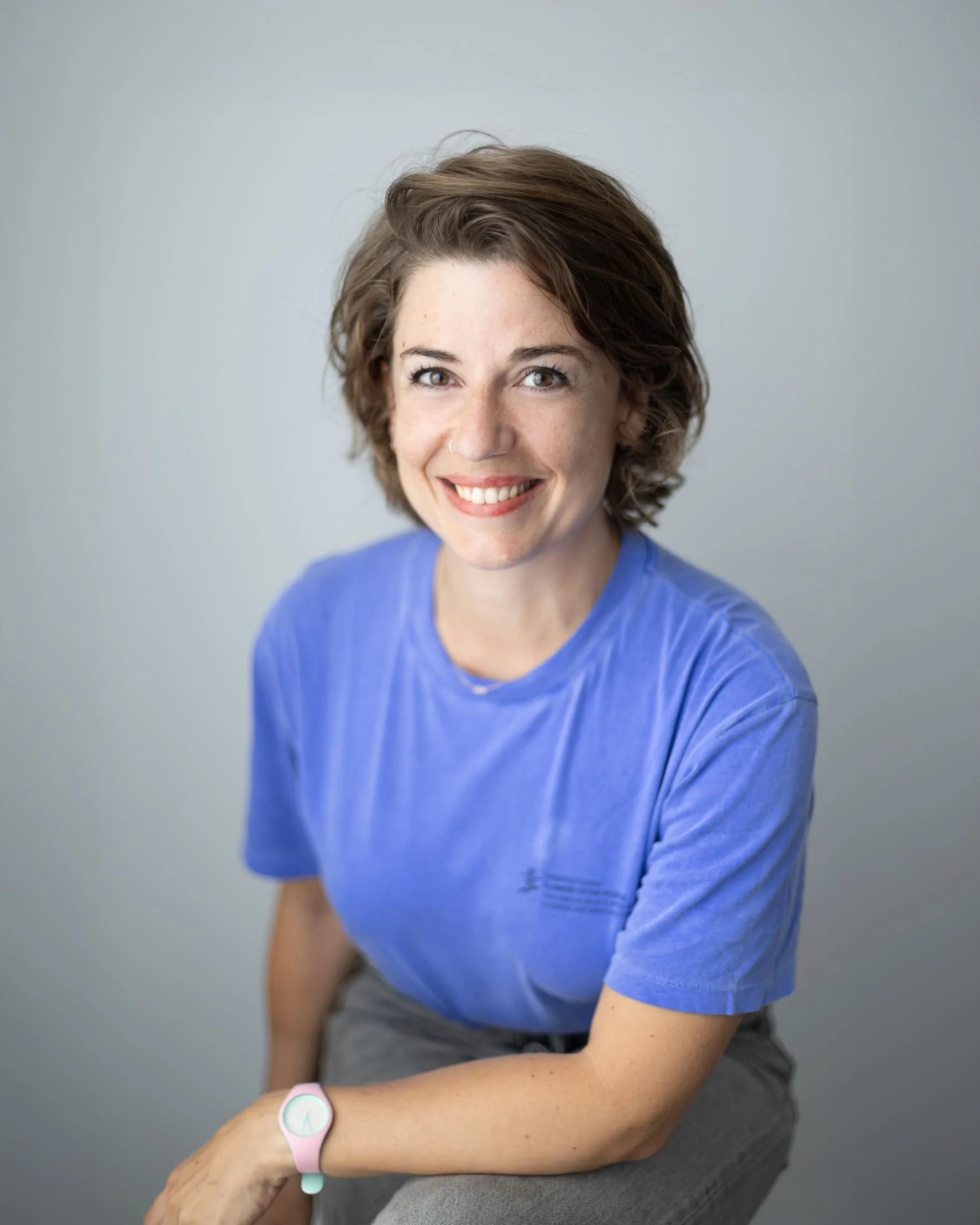 Jeune femme avec cheveux courts, souriant, portant un t-shirt bleu, assise, avec un bracelet intelligent rose et blanc, contre un fond gris clair.