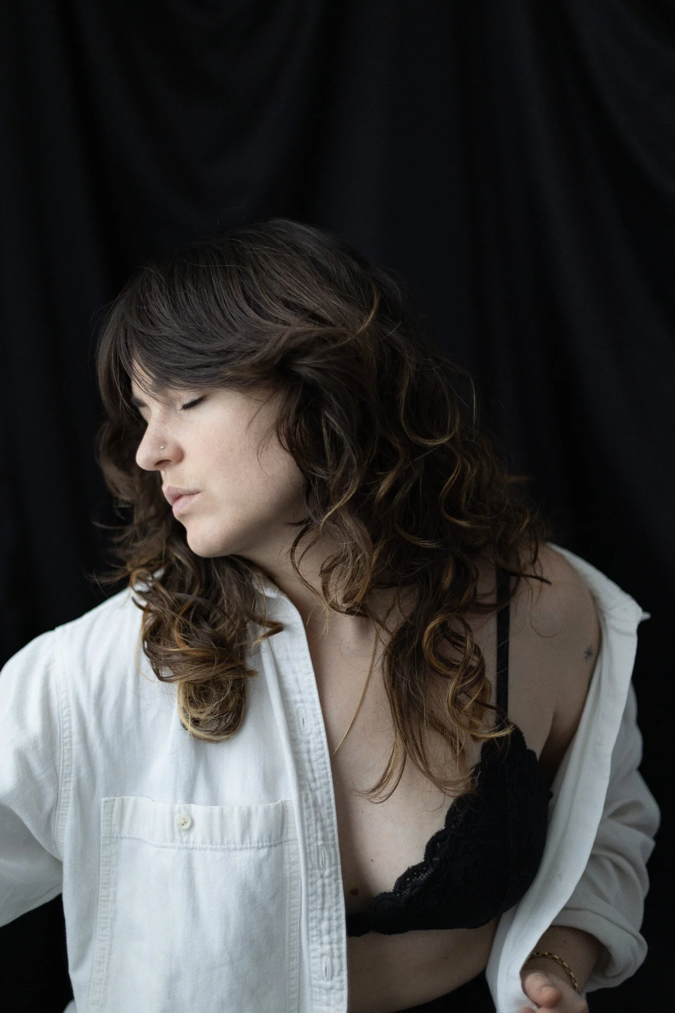 Une femme aux cheveux bruns bouclés, portant une chemise blanche, une lingerie noire et un collier doré, contre un fond noir.