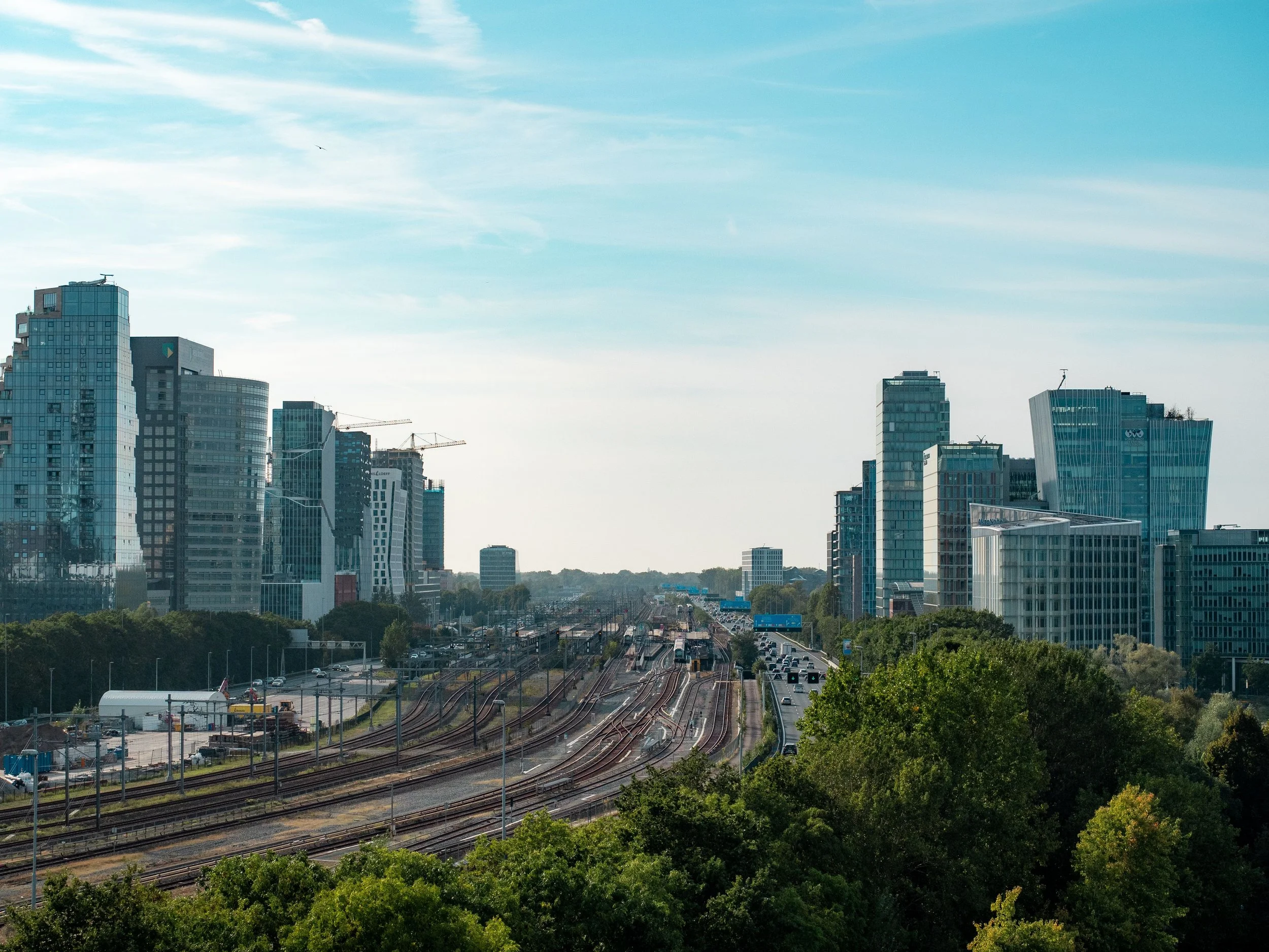 Hoge moderne kantoorgebouwen in een stedelijk landschap met sporen en verkeer op de voorgrond, onder een deels bewolkte lucht.