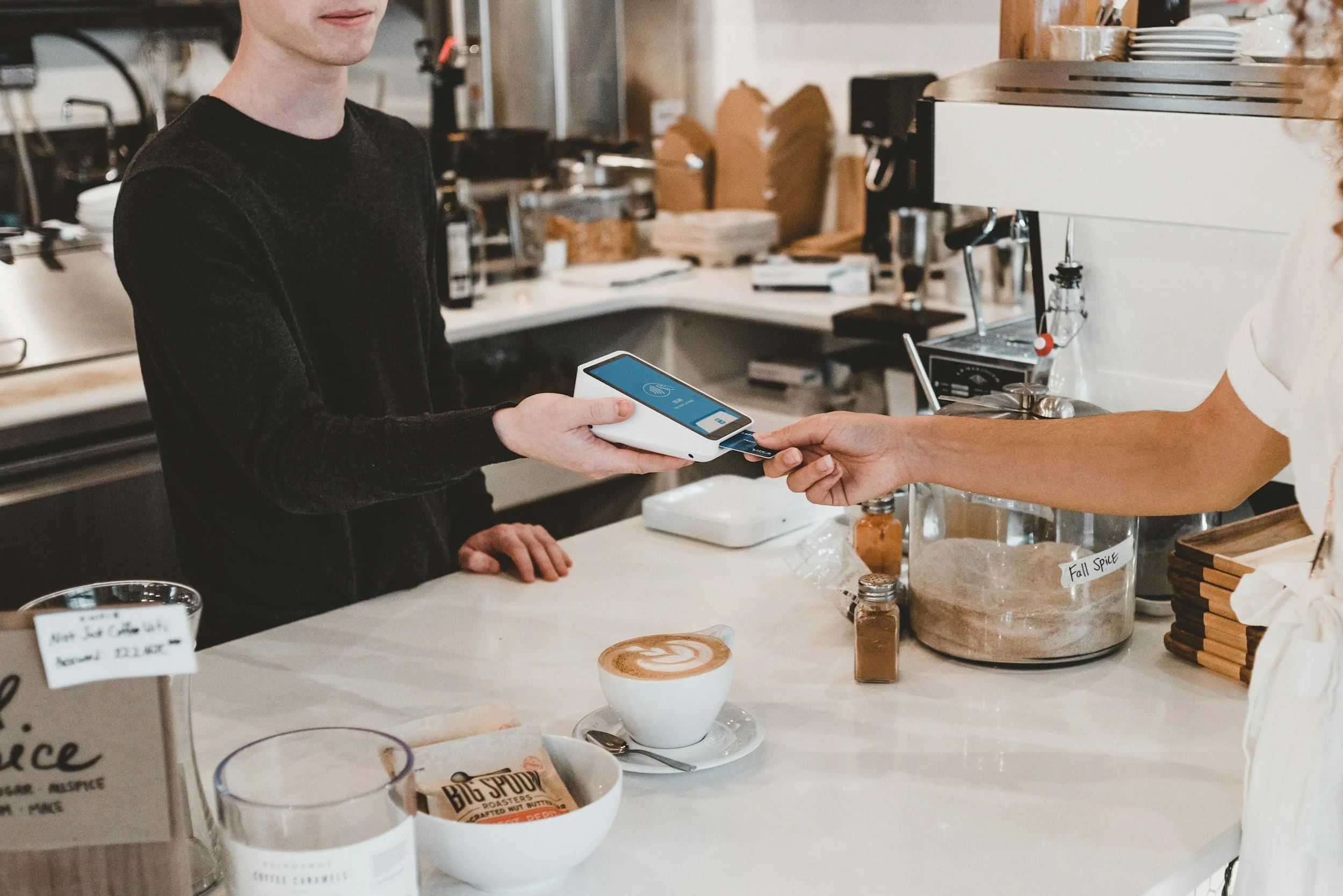 Man betaalt met een creditcard bij een barista in een koffiebar, met latte art koffie en ontbijtproducten op de toonbank.