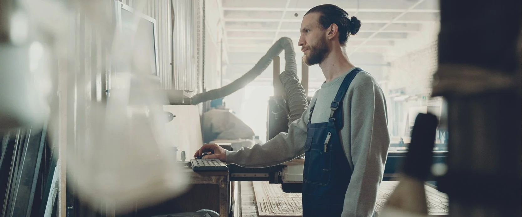 Een man met een paardenstaart en een baard, werkend in een werkplaats met machines, terwijl hij naar een scherm kijkt.