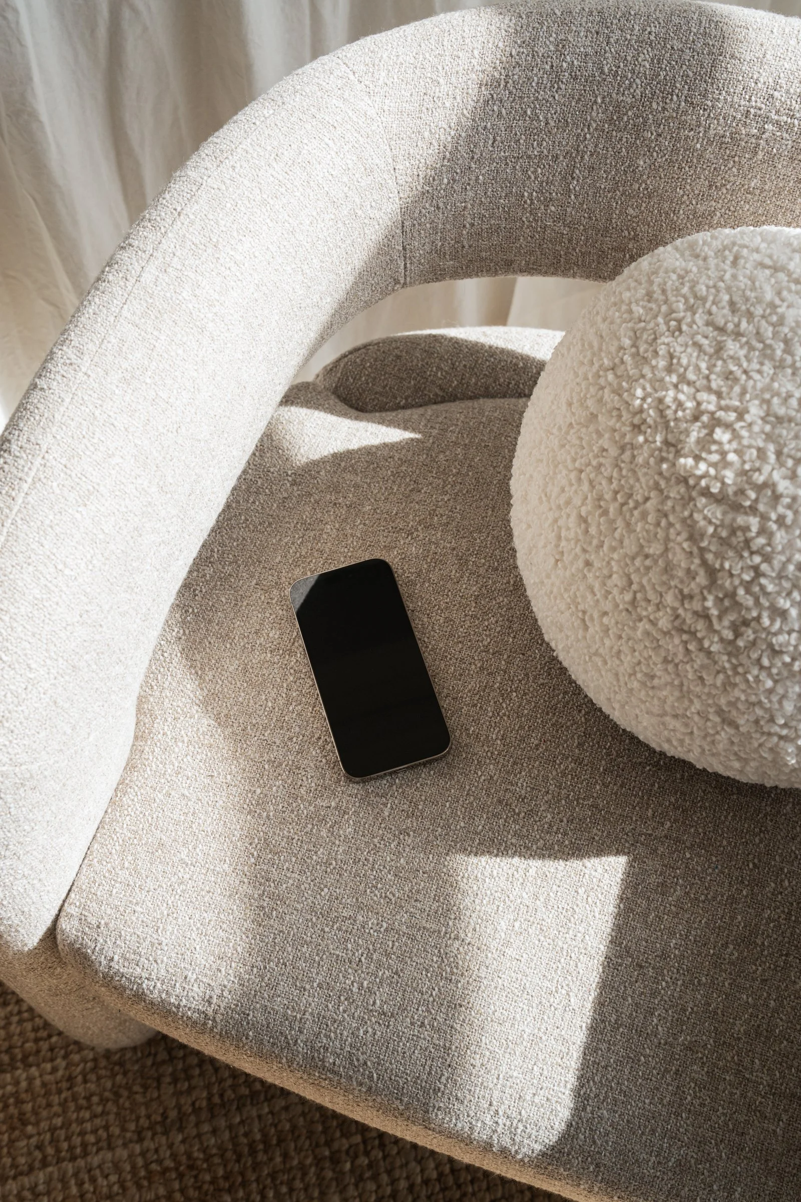 Een beige fauteuil met een zwarte smartphone en een ronde, witte zachte kussens.