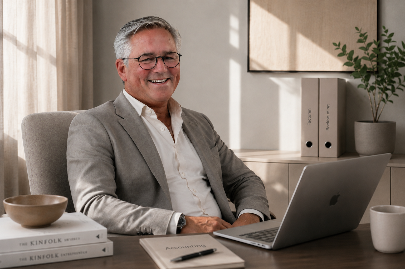 Een glimlachende man in een beige pak zit aan een bureau met een laptop, boeken en een plant op de achtergrond.