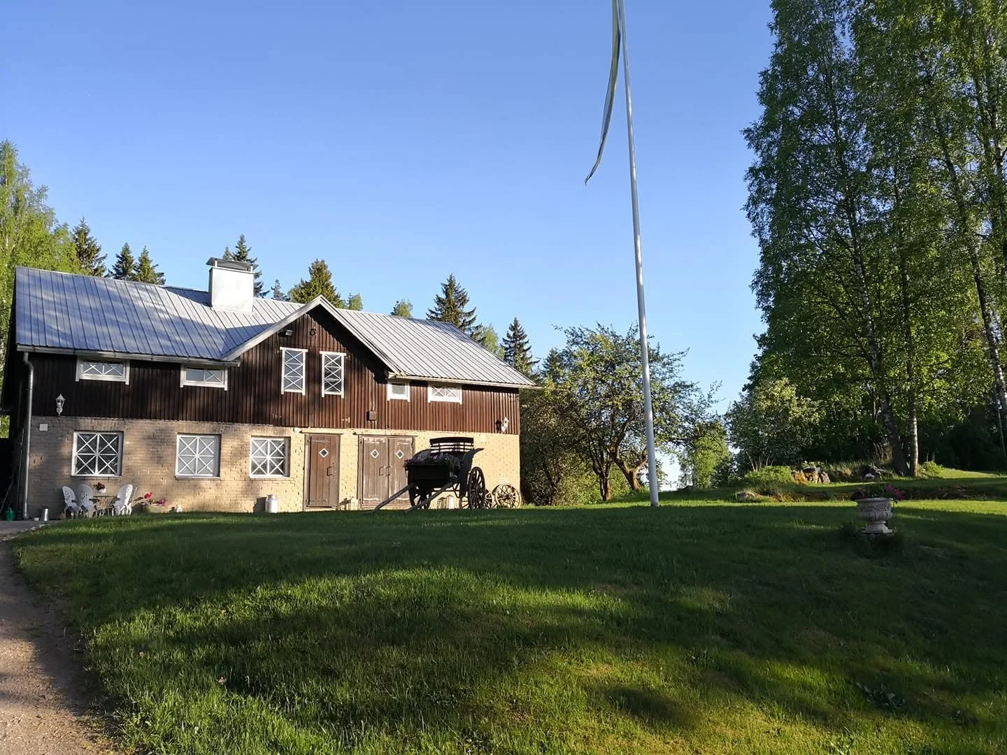 Metsäkyläinen talo, jossa on katto ja päädyt, ympärillä vihreitä puita ja nurmikkoa, sekä vanha kärry etualalla.
