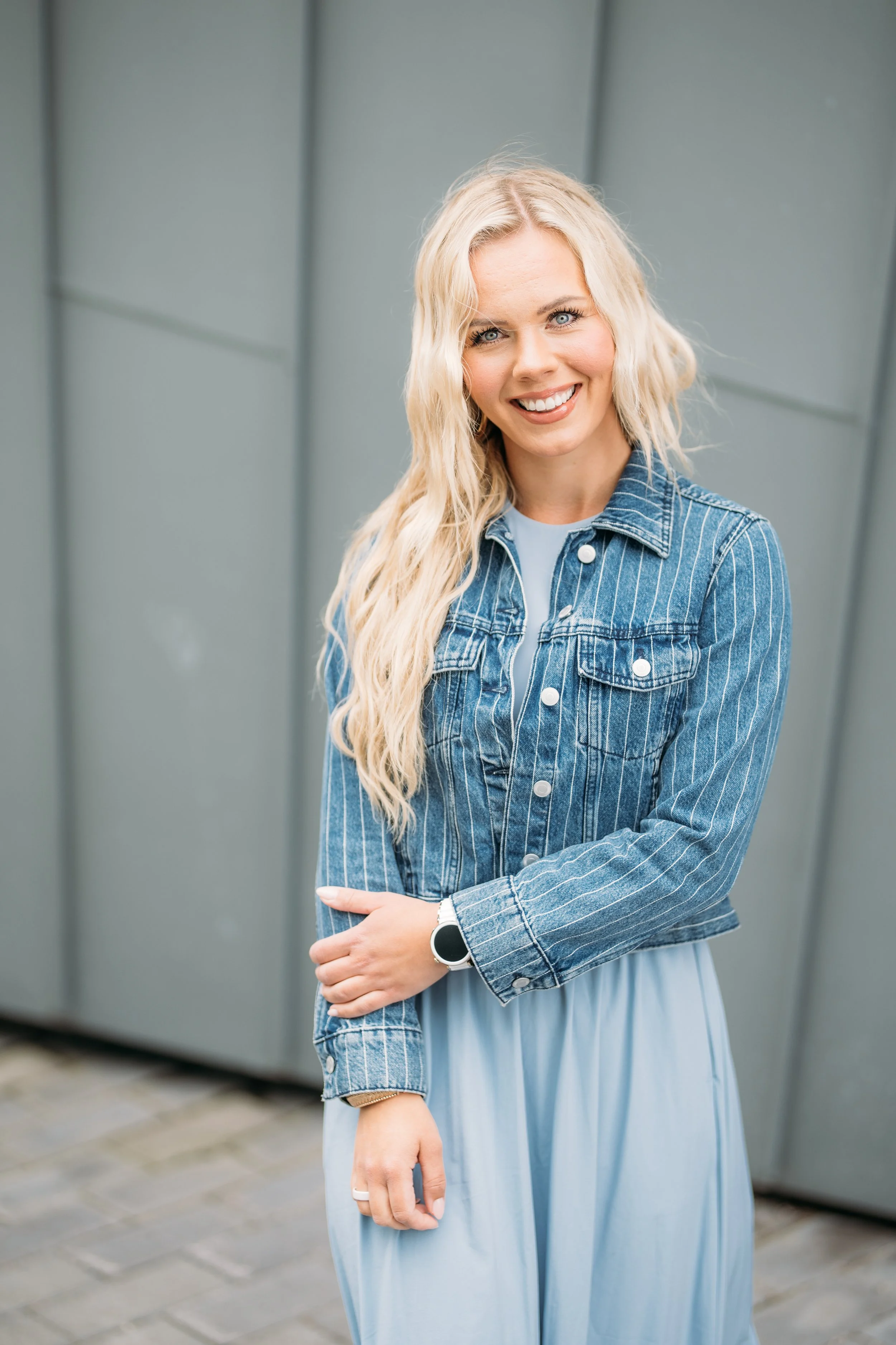 Hymyilevä nuori nainen tyylikkäässä denim-jakussa ja valkoisessa mekossa seisoo ulkona taustalla harmaat seinäpaneelit.