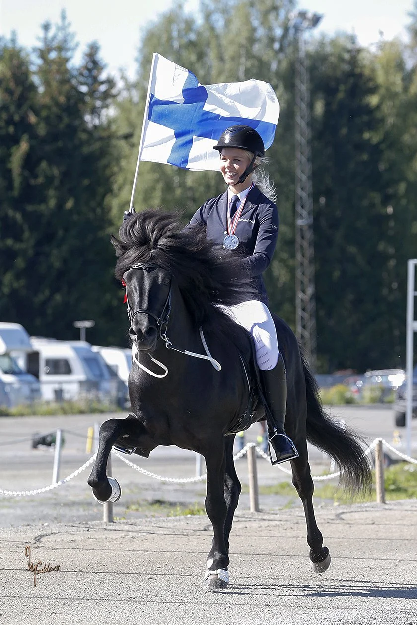 Nuori nainen ratsastaa mustalla hevosella, hänellä on kypärä, mitali kaulassa ja Suomen lippu kädessään, taustalla puita ja autoja.