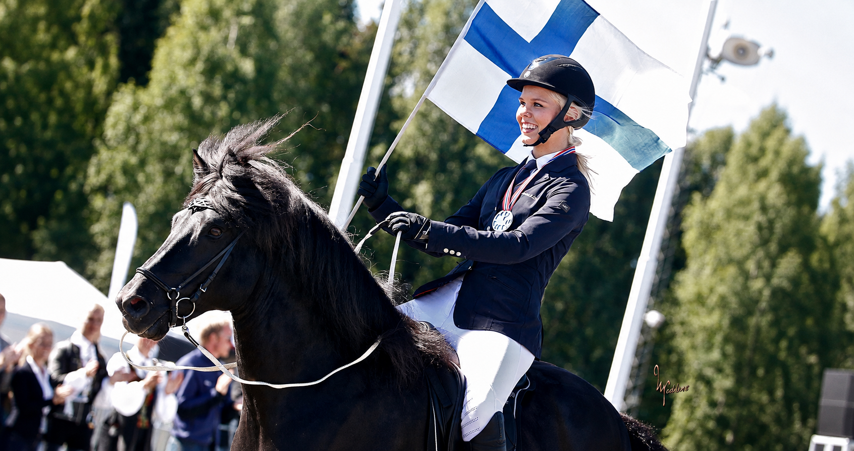 Nainen ratsastaa kilpailussa, pitää Suomen lippua, ja hänellä on pronssimitali, hän on pukeutunut urheiluvaatteisiin ja kypärään, taustalla yleisö ja vihreitä puita.