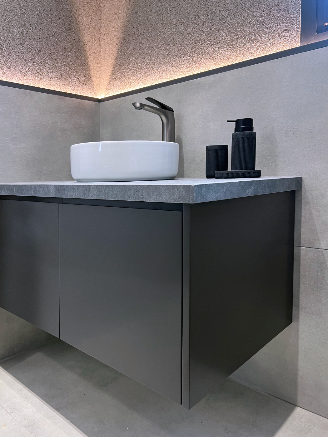 Modern bathroom vanity with a vessel sink, black soap dispenser, and toothbrush holder against a textured wall with ambient lighting.