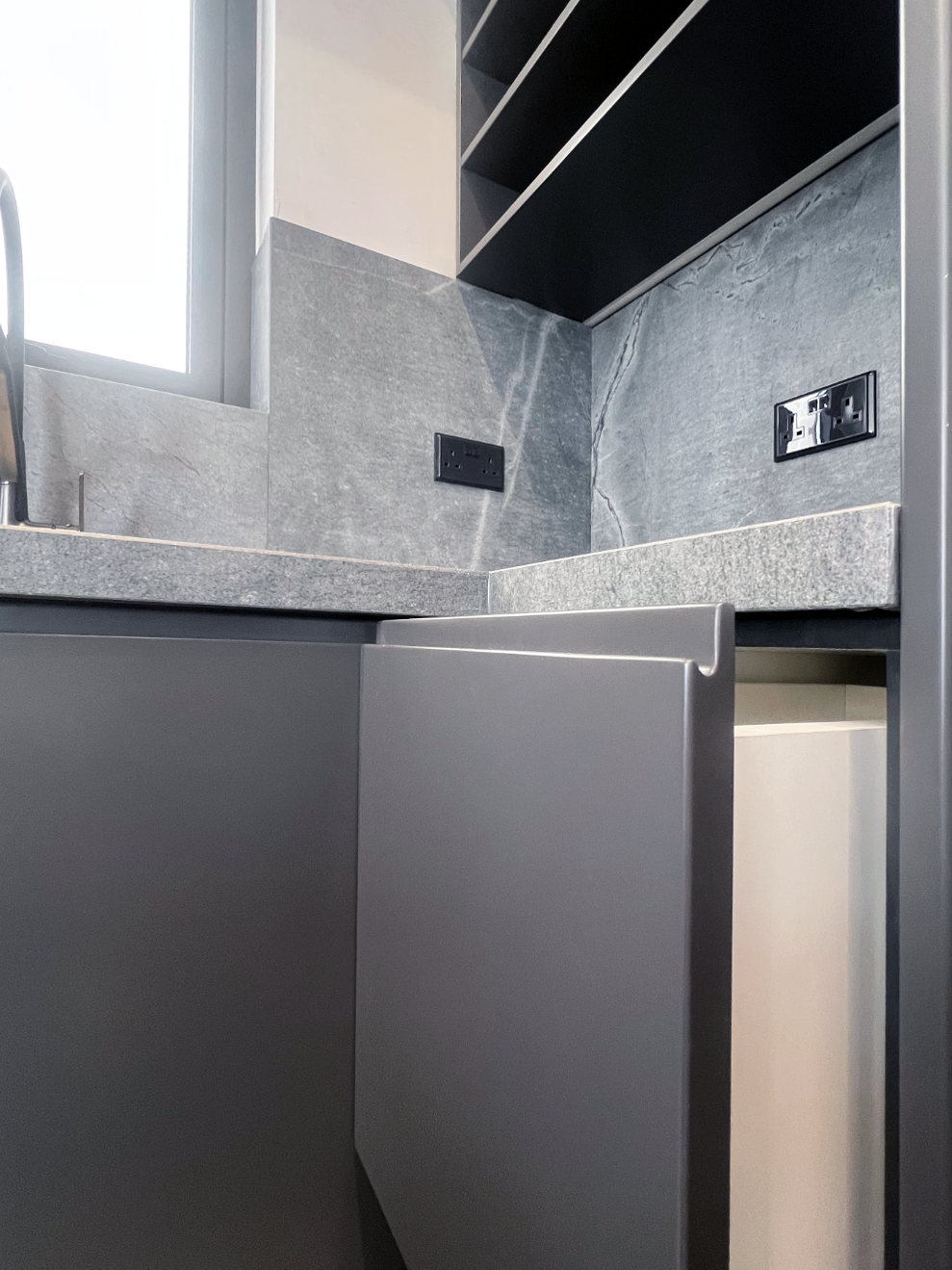 Close-up view of a modern kitchen counter with gray cabinetry, gray stone surface, and multiple electrical outlets on the wall, with a window letting in natural light.