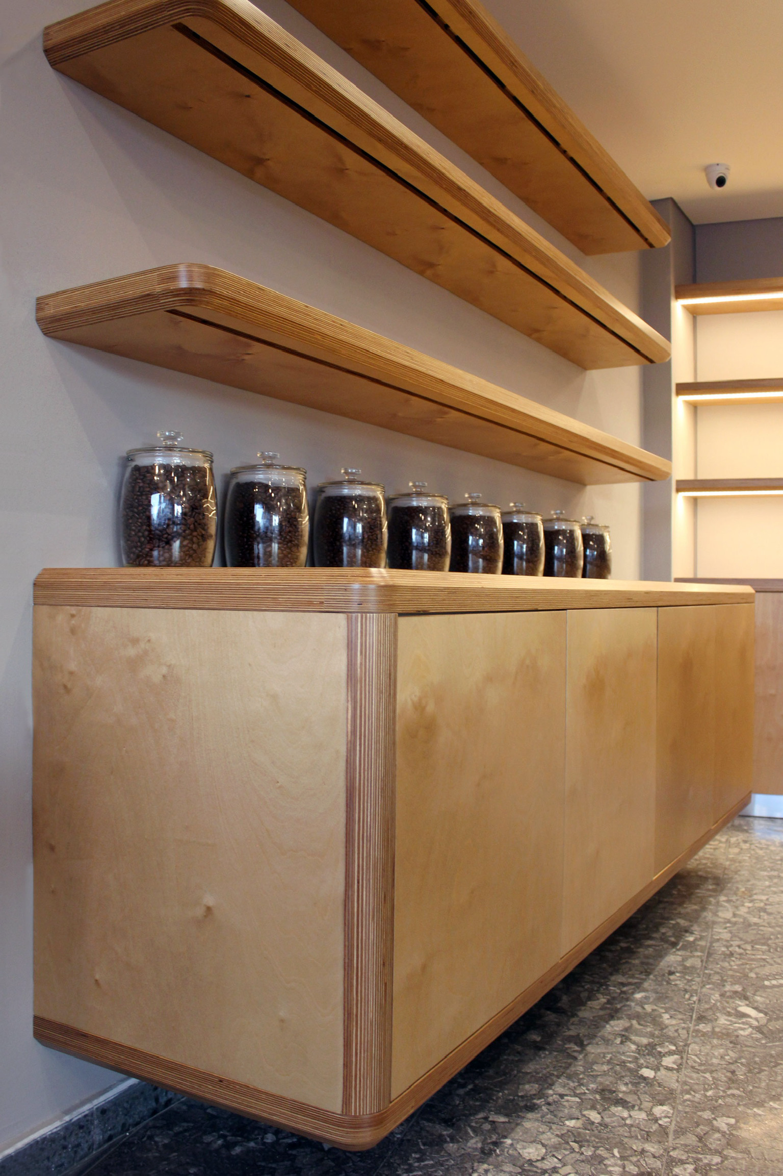 Wooden shelves mounted on a wall above a wooden cabinet with glass jars of coffee beans on top. The shelves are light-colored wood with rounded edges and black slots for brackets.
