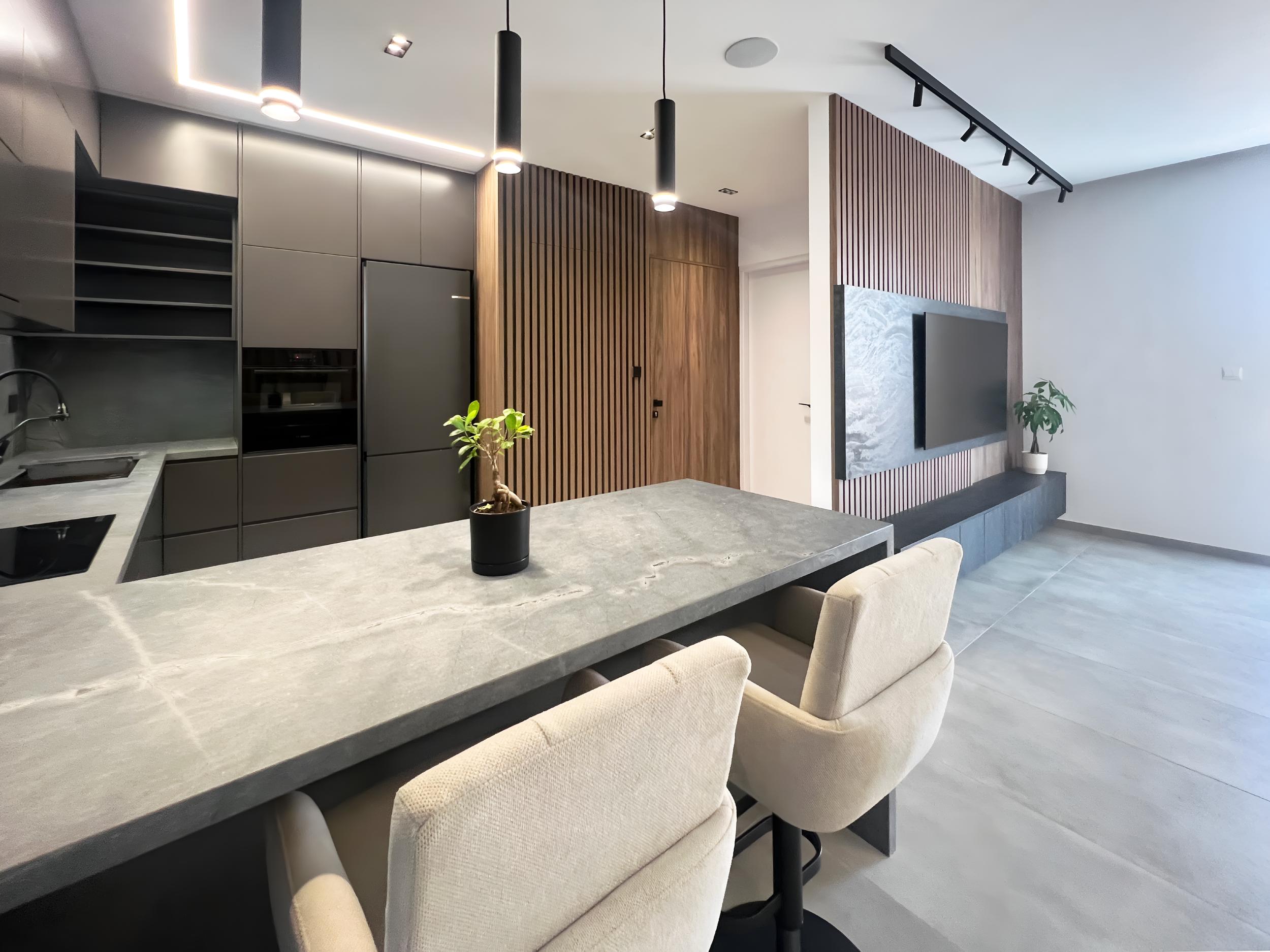 Modern living room with a gray kitchen island and beige upholstered chairs around it. In the background, there are dark gray cabinets, wooden wall panels, a mounted flat-screen TV on a wood-paneled wall, and potted plants. The room features ceiling l