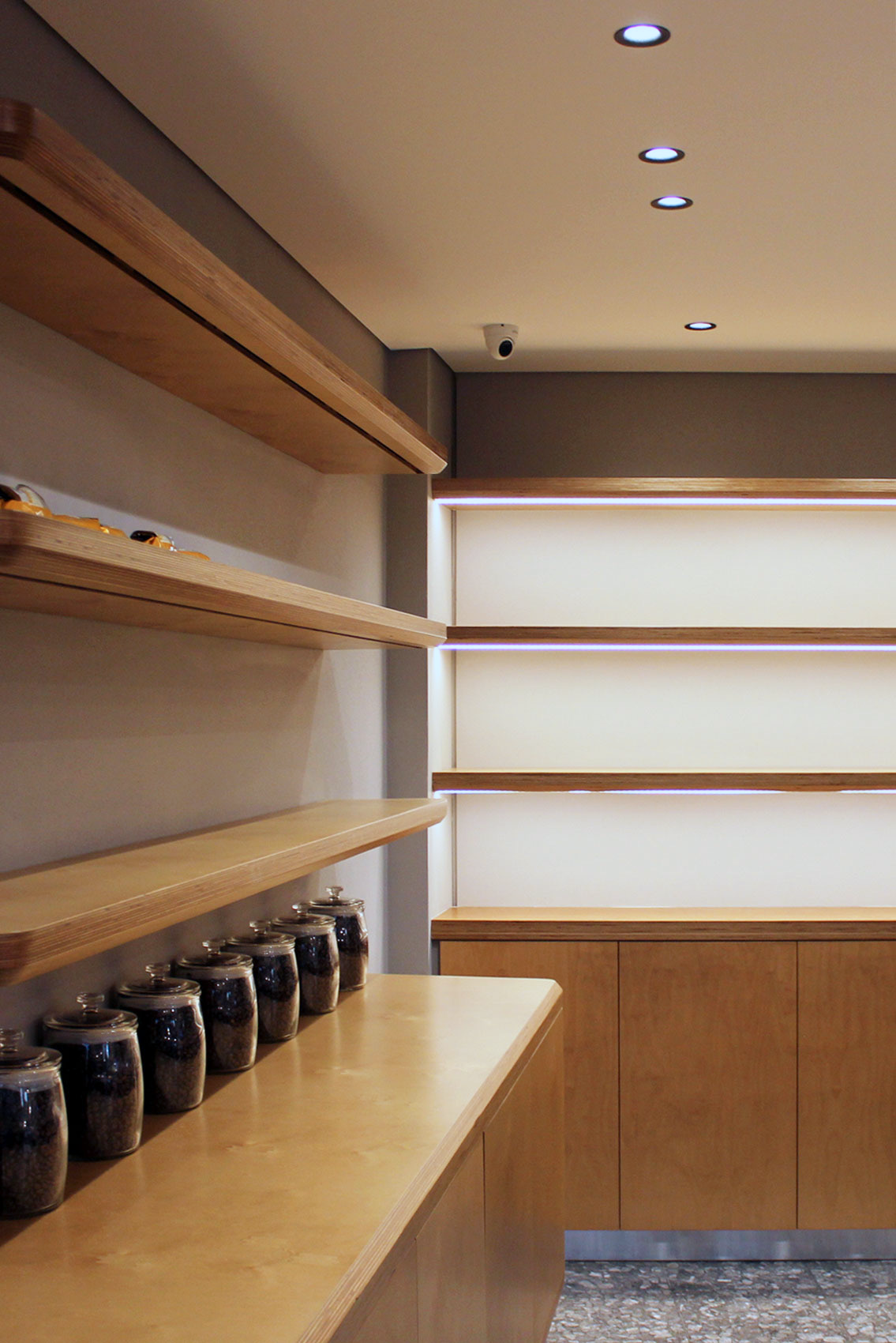 Empty wooden shelves and cabinet in a modern store or cafe. Glass jars with dark contents line the lower shelf. Ceiling with recessed lighting and a security camera.