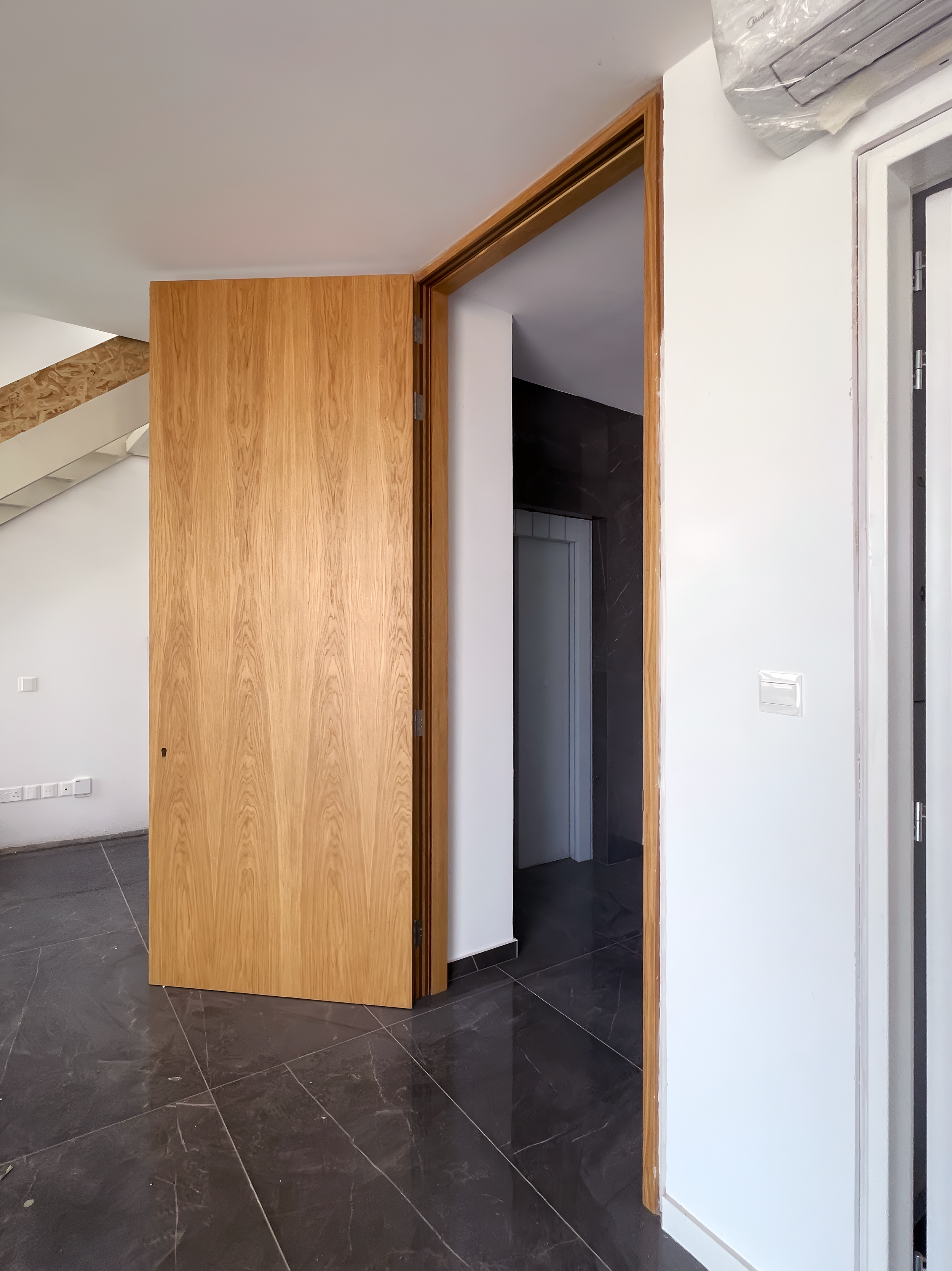 Interior of a room with a partially open wooden door, white walls, black marble floors, and a staircase with wooden railing on the left side. Visible door frame and electrical switches.
