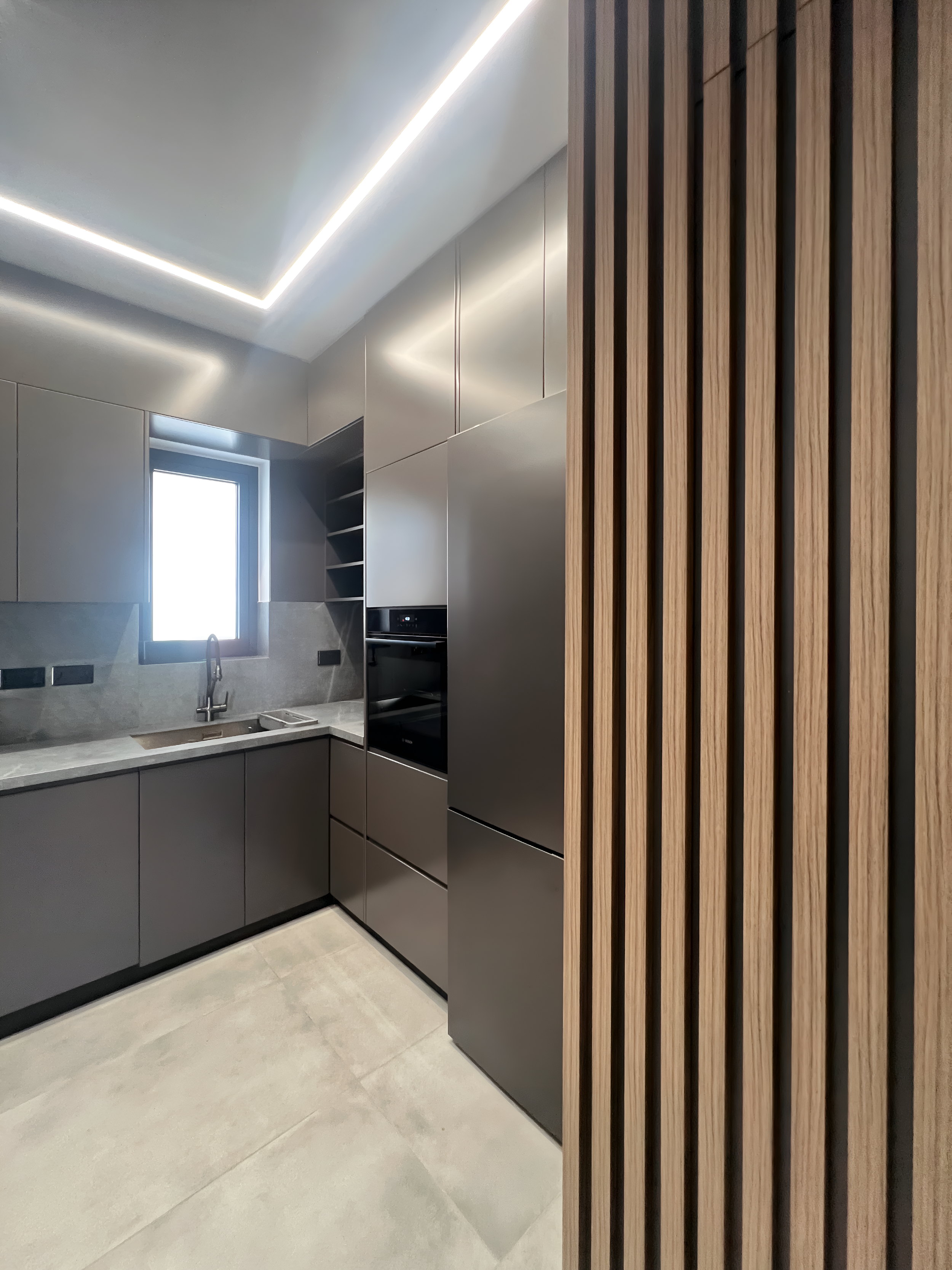 Modern kitchen with gray cabinets, a marble countertop, and a stainless steel sink. A window above the sink lets in natural light, and a wooden slat divider or wall is visible on the right side.