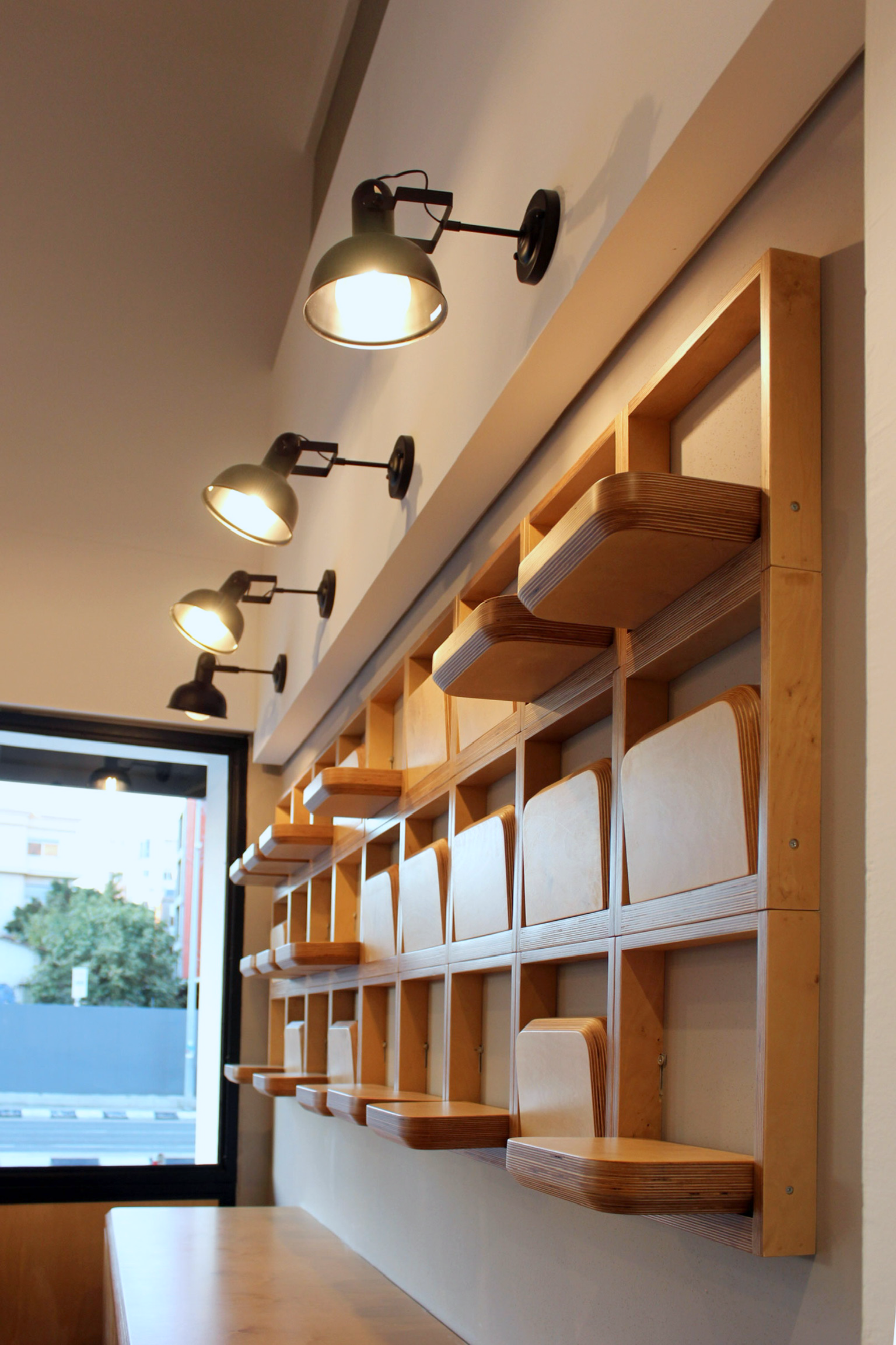 Interior of a room with a wooden shelving unit mounted on the wall, featuring multiple individual cubbies, with black wall-mounted directional lamps above and a window with a view of trees and buildings outside.