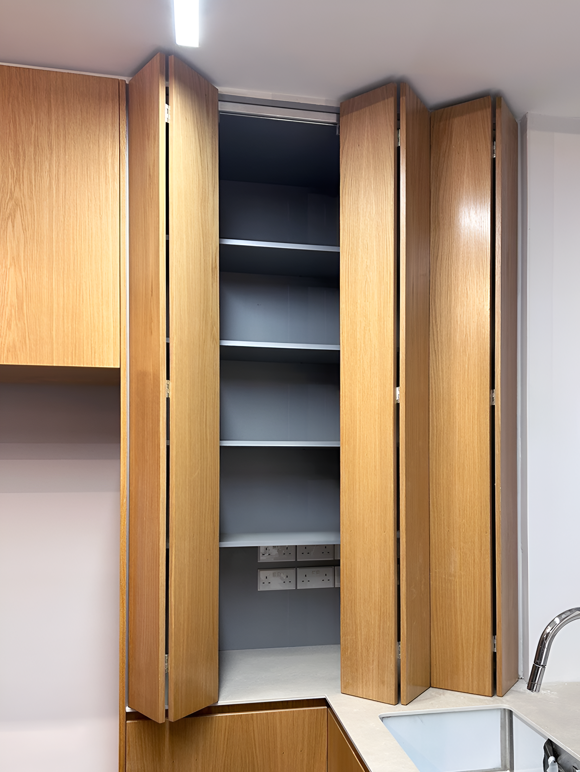 Empty kitchen cabinets with wooden doors open above a beige countertop, a modern kitchen sink, and electrical outlets on a gray wall.
