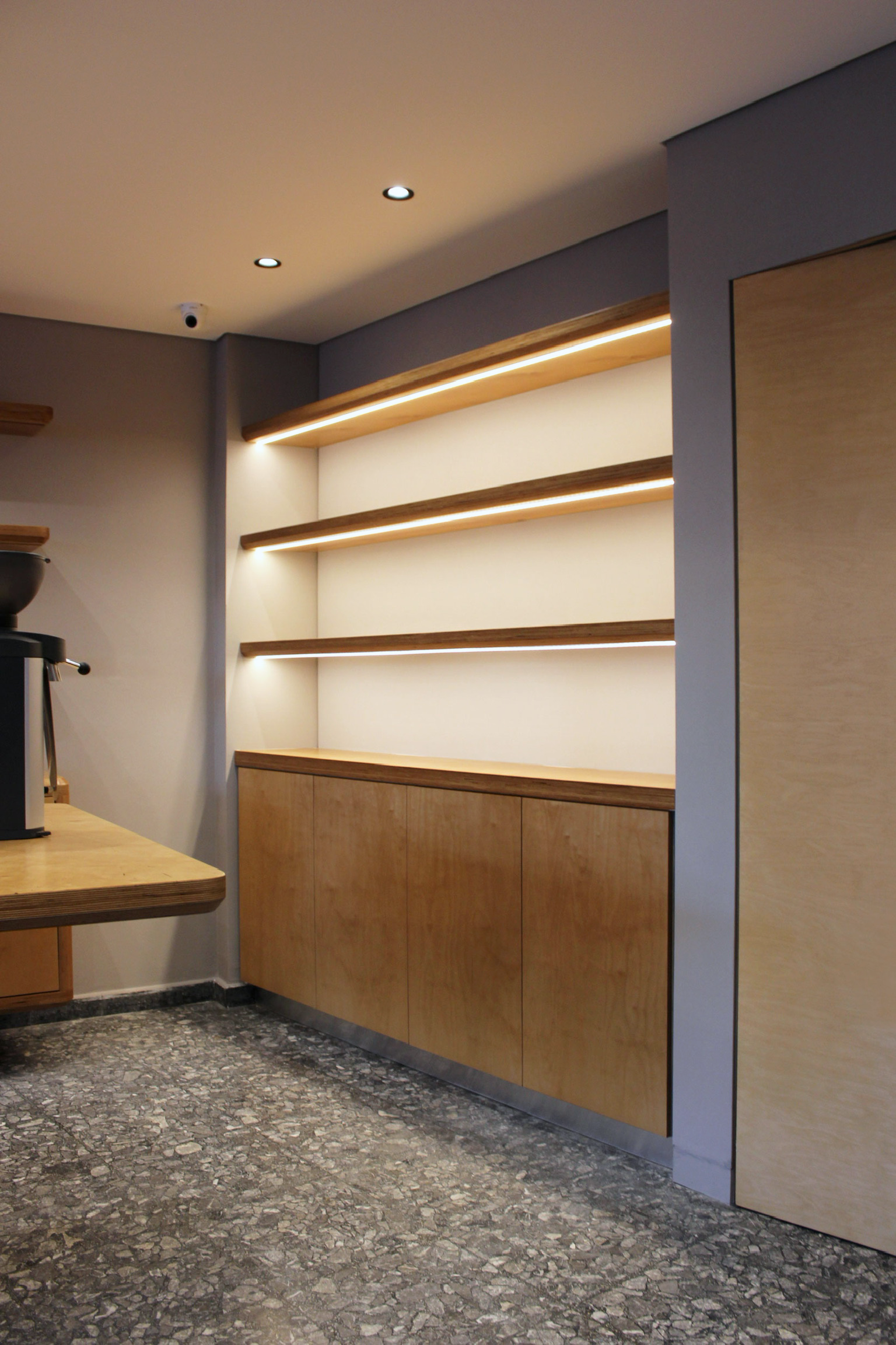 Inside a modern room with wooden shelves with LED lighting, a wooden cabinet, and a coffee machine on a wooden table. The floor is covered with dark stone tiles, and the ceiling has three small round recessed lights and a security camera.