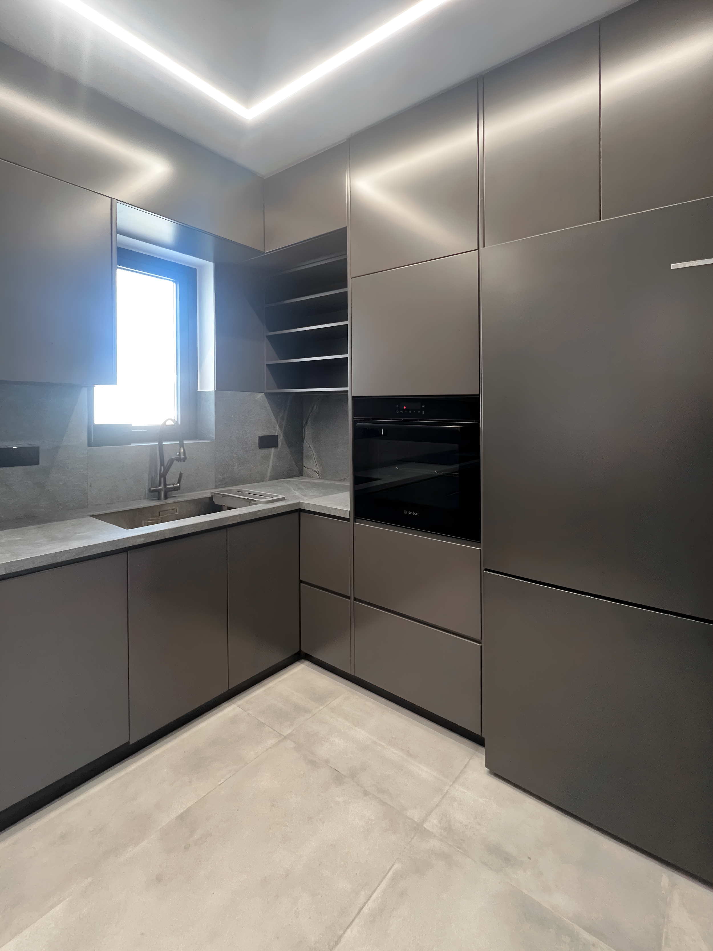 Modern kitchen with grey cabinetry, a window, sink, and built-in oven, with beige tiled floor.