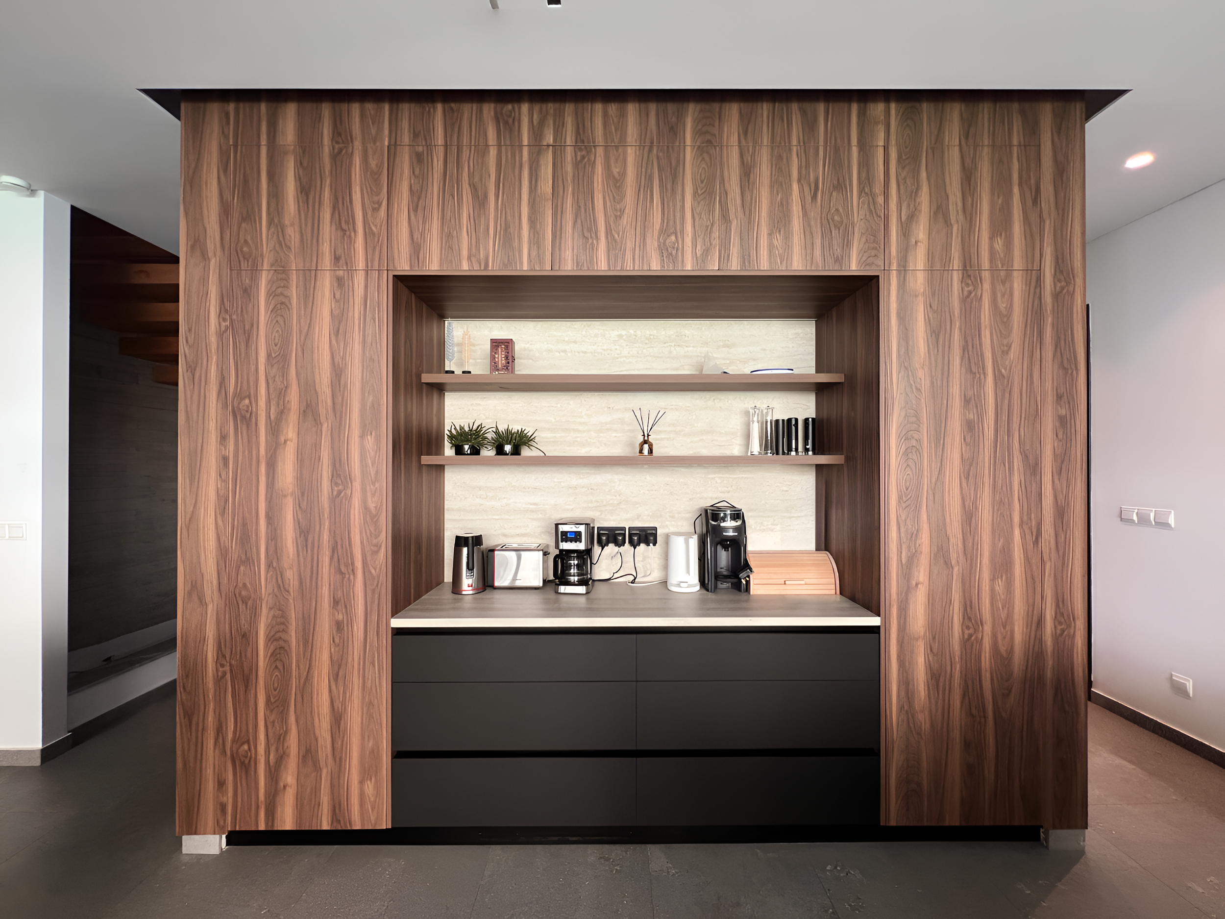 A kitchen with a wooden wall and built-in shelves holding plants, books, and decorative items, with kitchen appliances and a countertop.