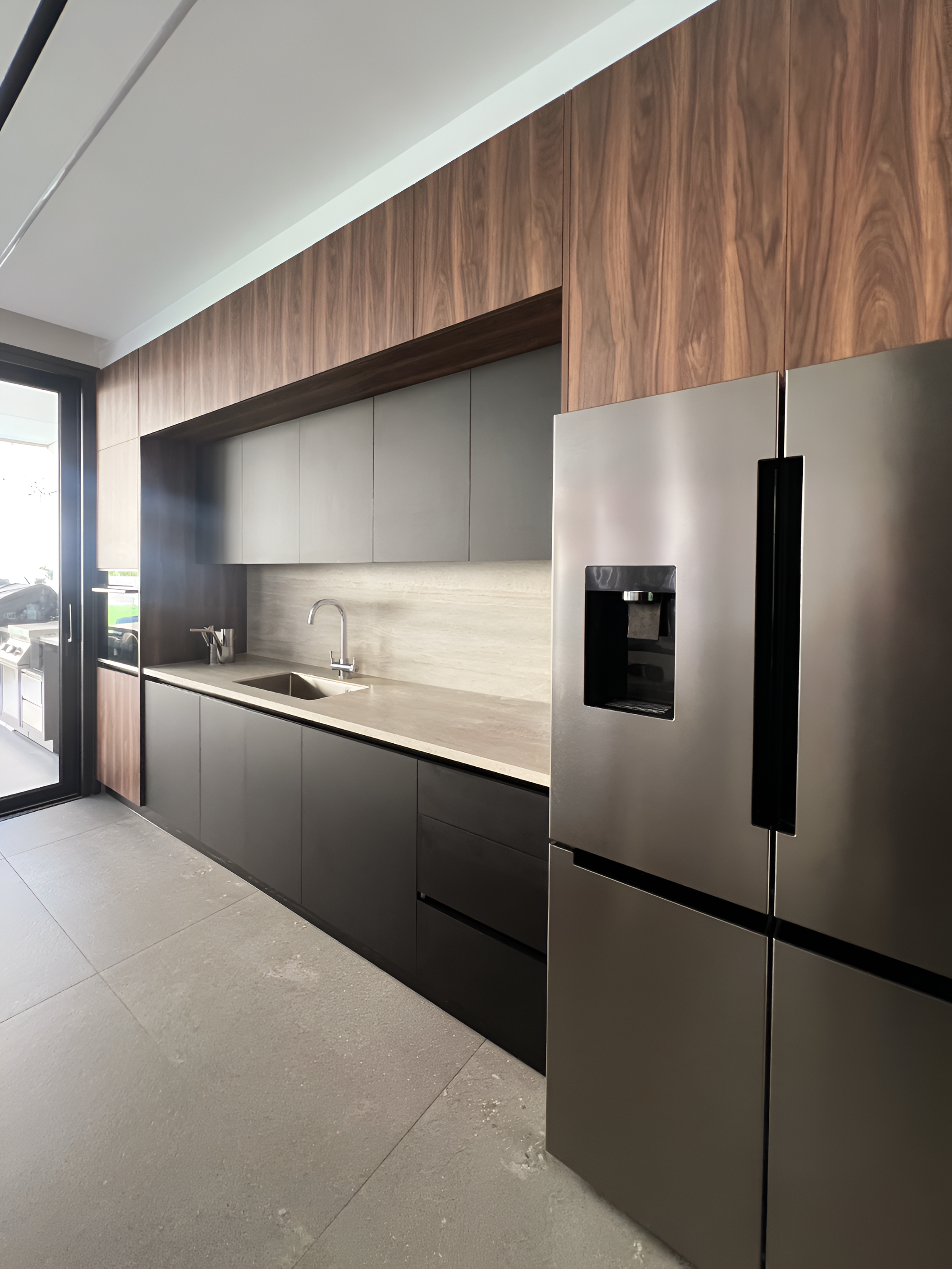 Modern kitchen with wooden upper cabinets, gray lower cabinets, stainless steel refrigerator, beige countertop, and a sink. The kitchen has large windows with sliding glass doors leading outside.
