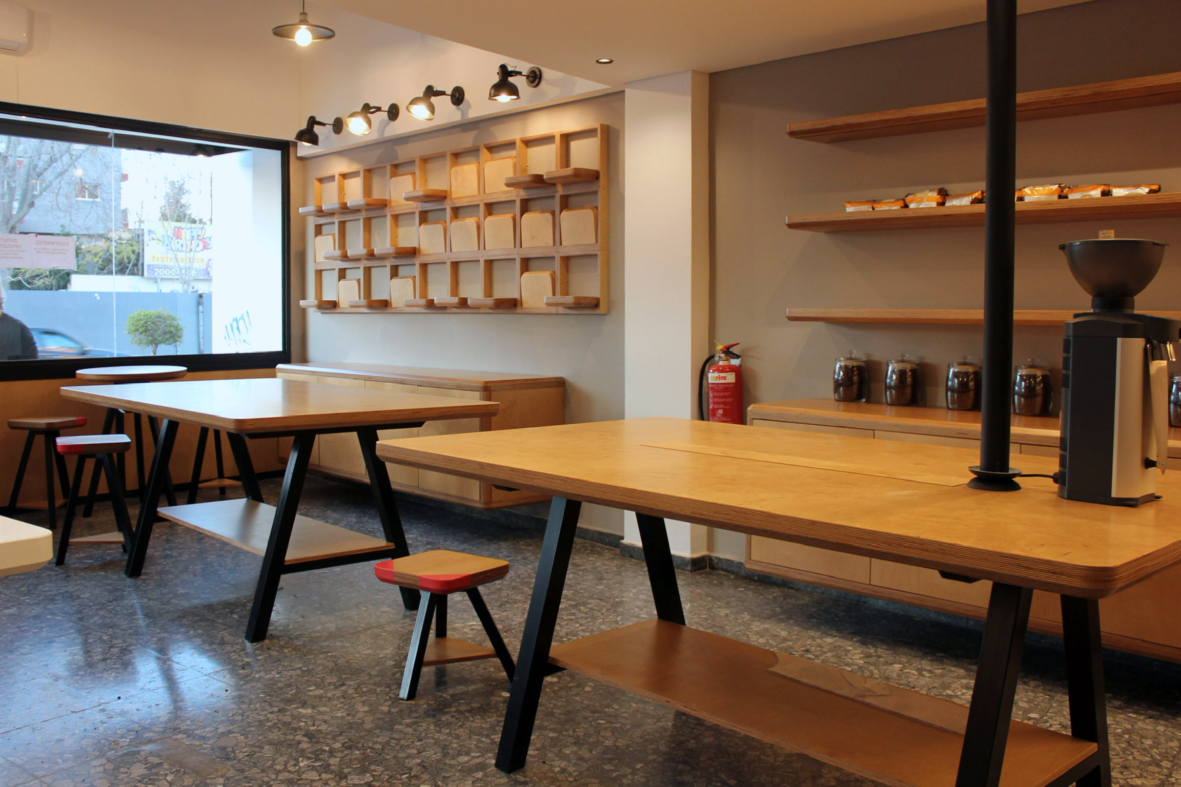 Interior of a modern cafe or restaurant with wooden tables, black chairs, and wall-mounted wooden shelves. Bright natural light from large window, ceiling lights, and a fire extinguisher visible.