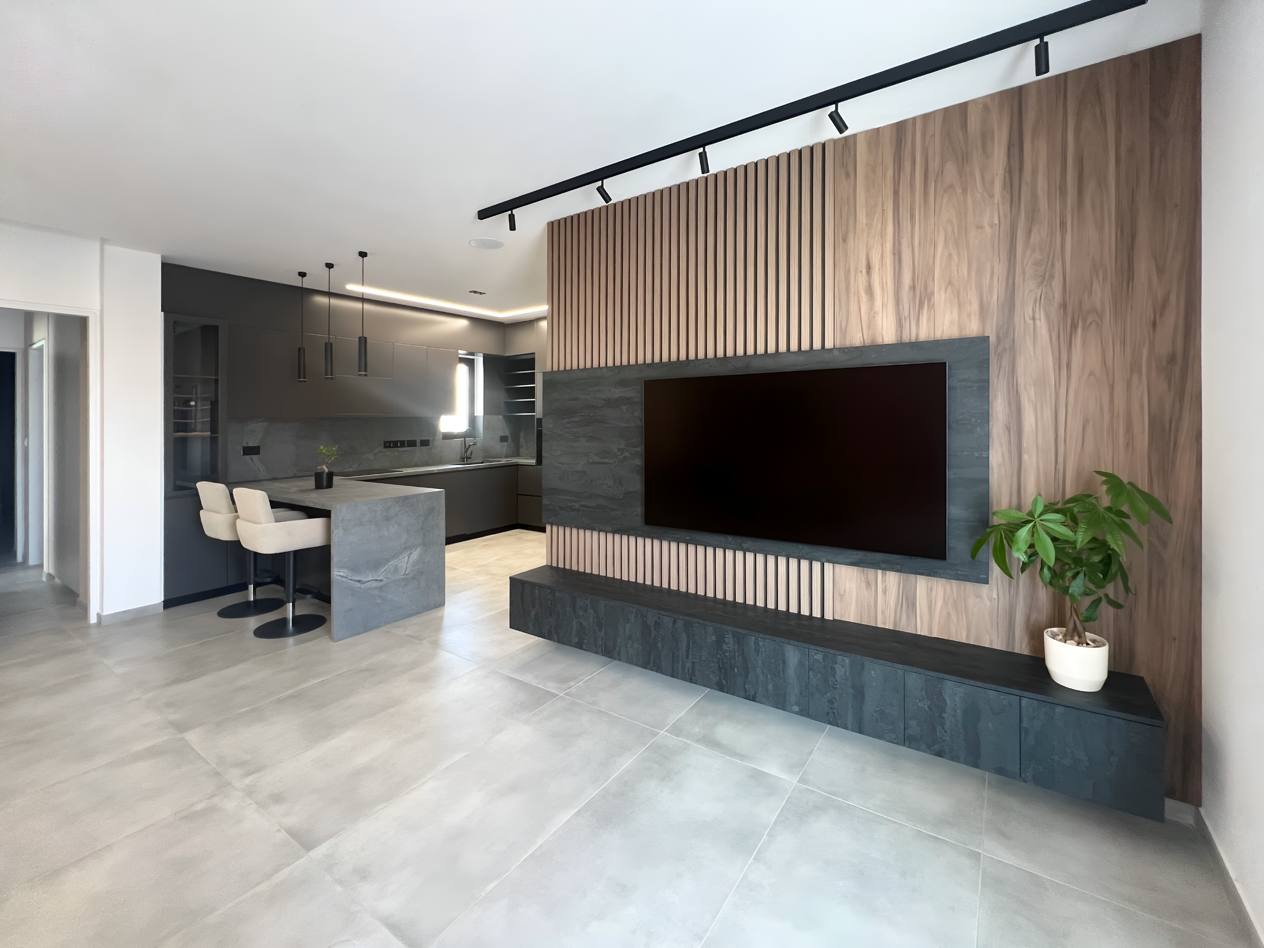 Modern living room with gray and wood accent walls, large gray tiles, and a mounted flat-screen TV. There is a small kitchen area with a marble-topped island, white upholstered bar stools, and pendant lighting. A potted plant is in the corner.