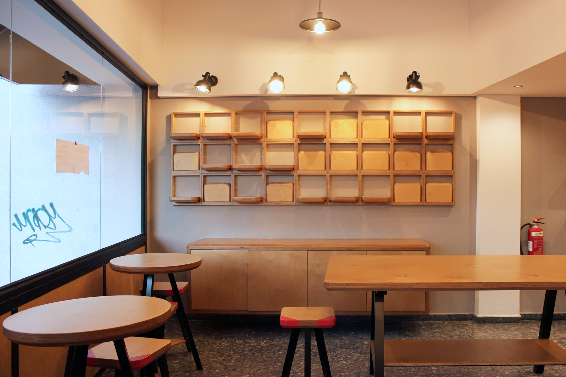 Interior of a modern cafe with wooden tables and chairs, a wall with empty wooden cubbies, and ceiling lights.