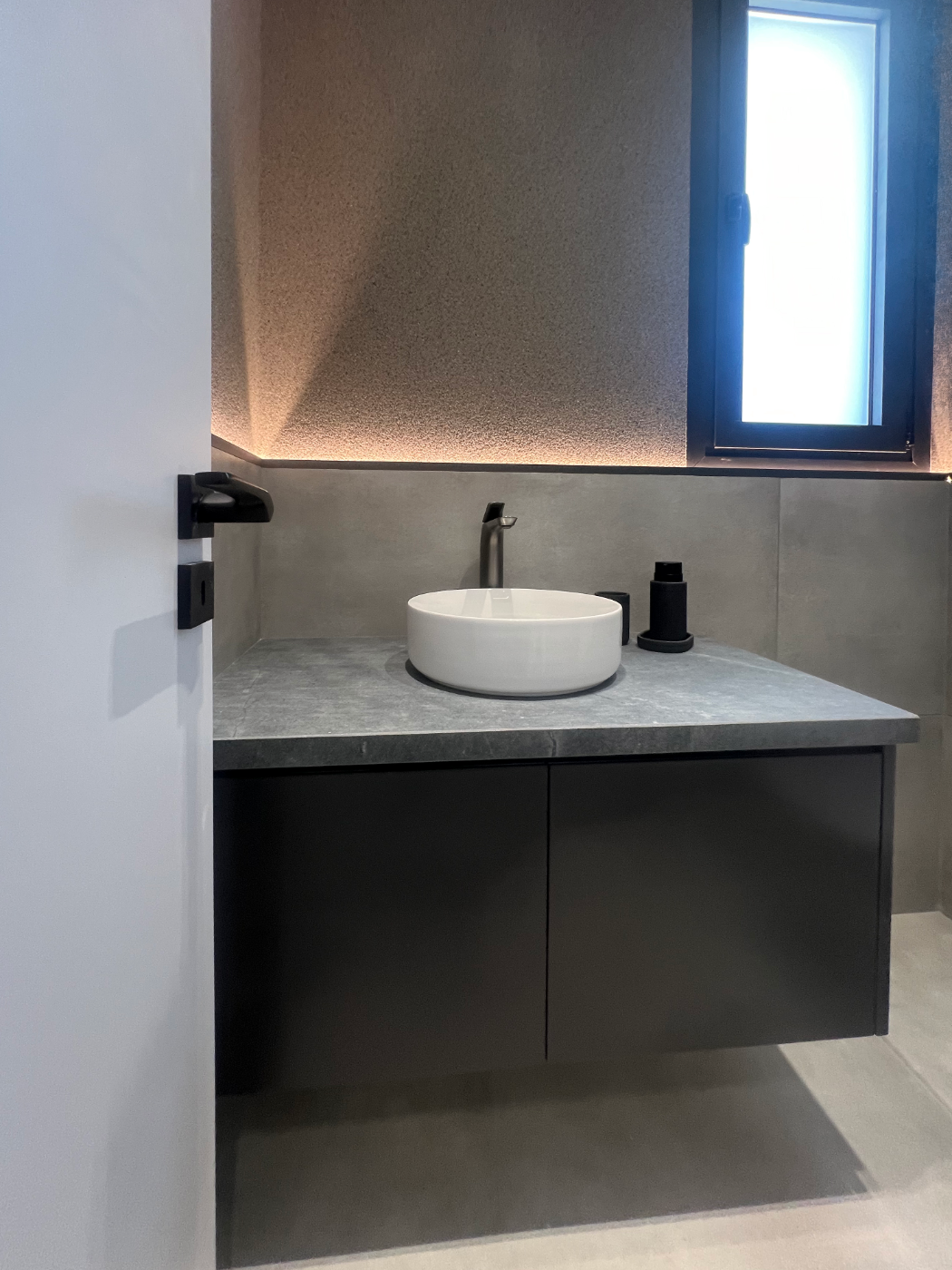 Modern bathroom with a small round white sink on a gray countertop, black soap dispenser, black cup, window with blue tint, and textured beige wall with ambient lighting.